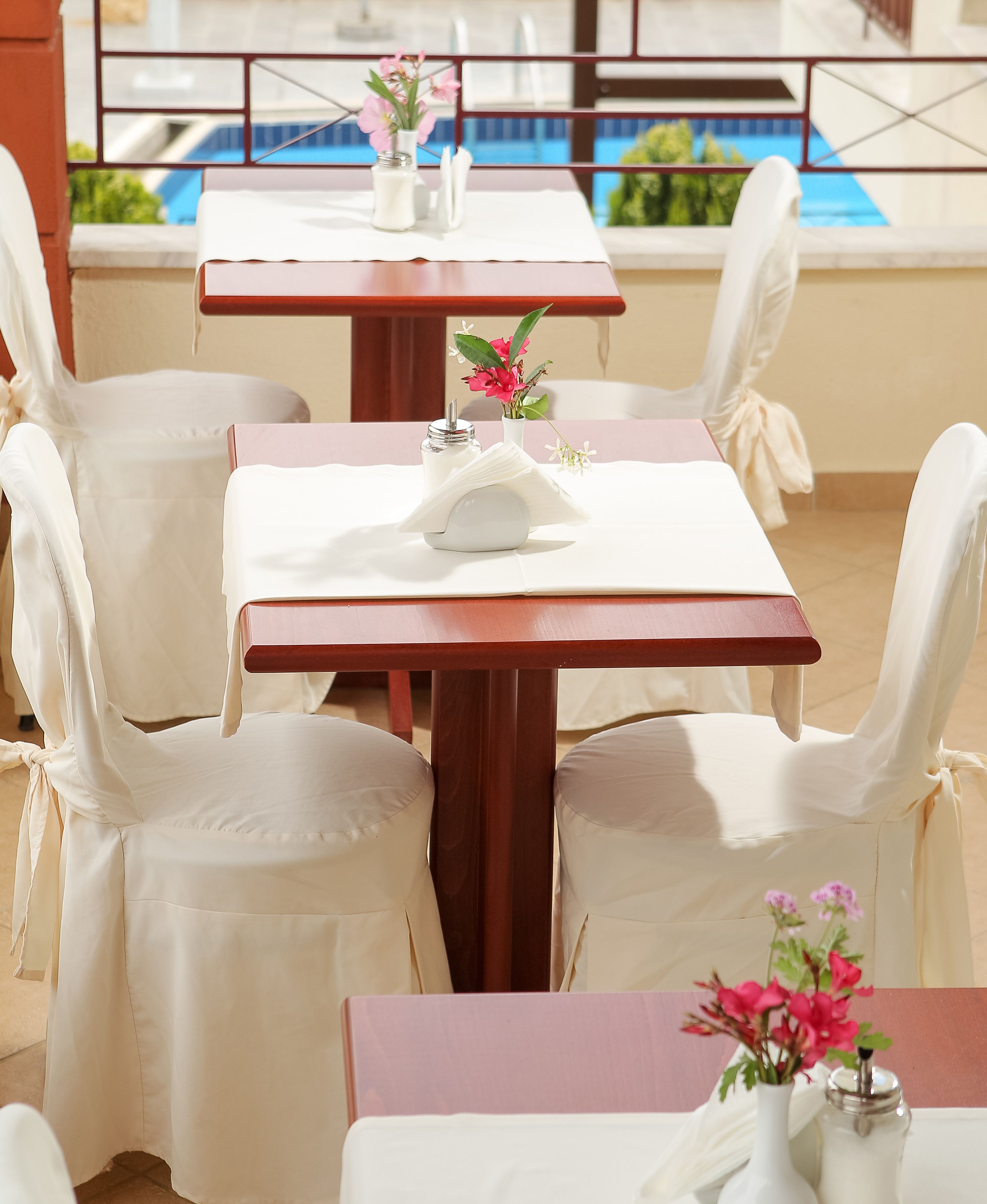 A dining area with tables and chairs, a vase with flowers, and a view of a pool.