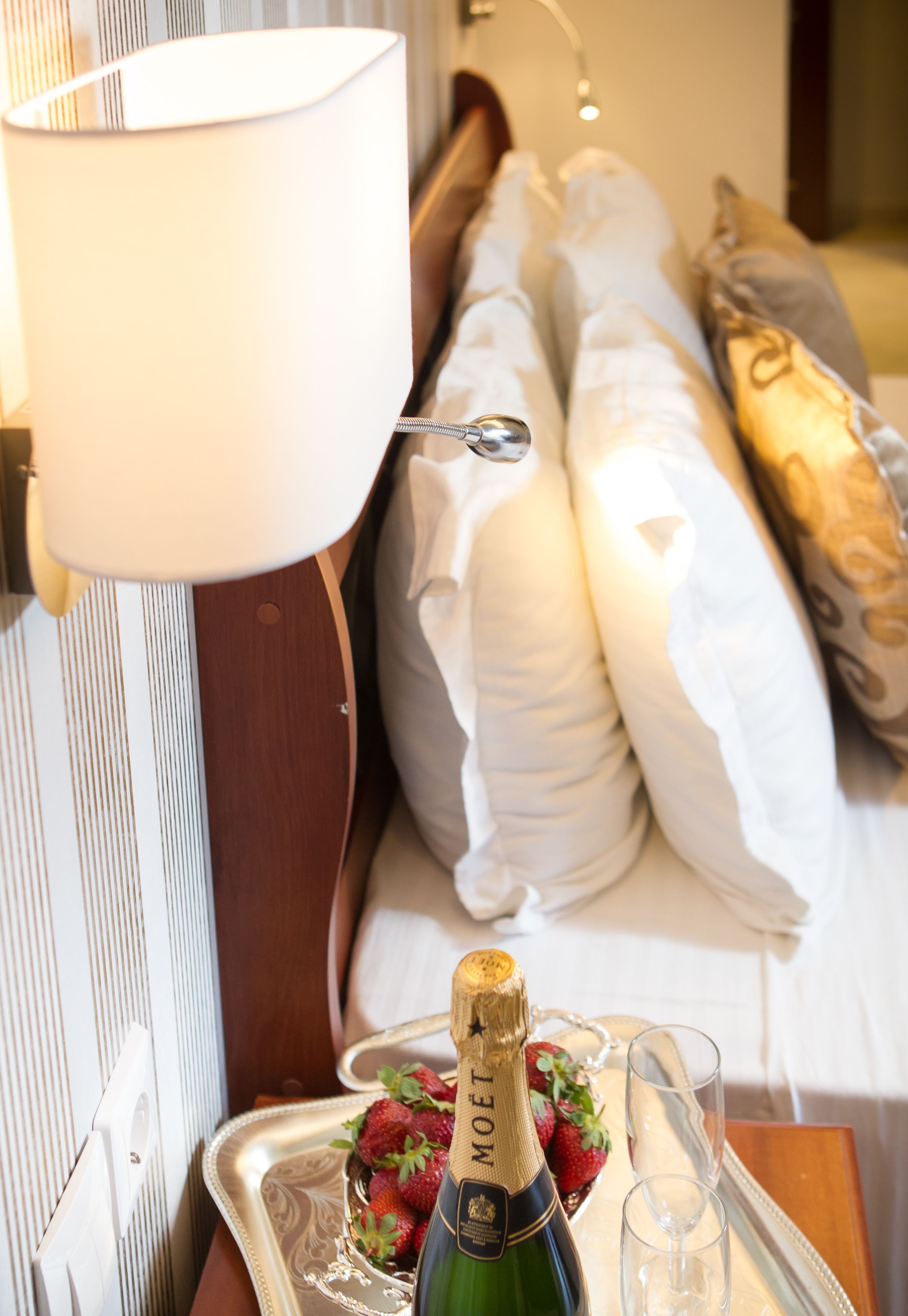 A hotel room with a bed, pillows, a bedside table with a bottle of champagne, strawberries, and glasses.