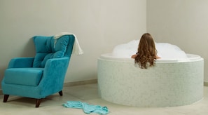 A room with a blue armchair, a bathtub with a person sitting in it, and a blue towel on the floor.