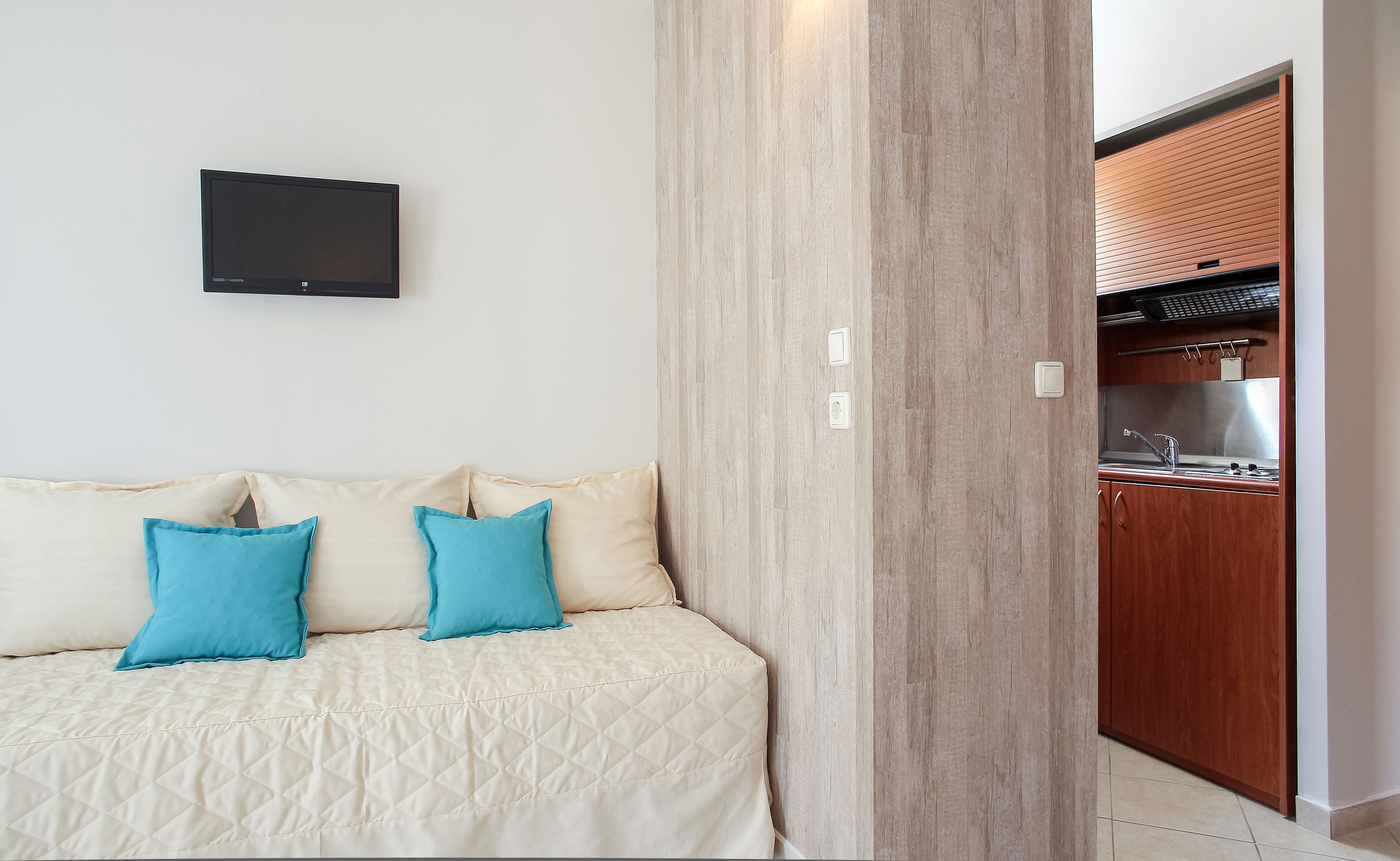 A bedroom with a bed, a TV mounted on the wall, and a wooden door leading to a kitchen.