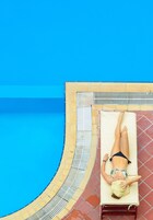 Una persona recostada en una tumbona junto a una piscina con suelo de baldosas y agua azul.