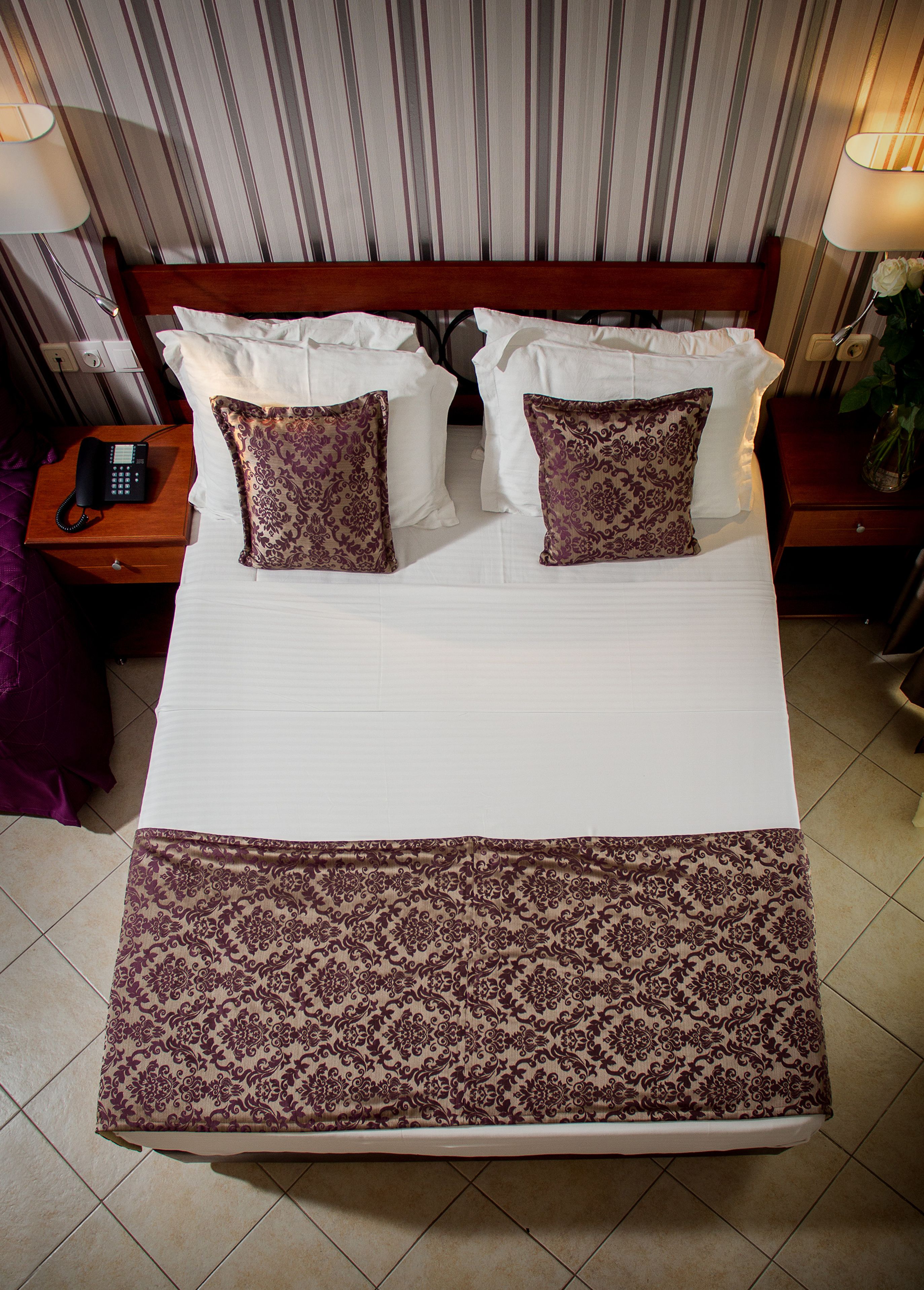 Cama doble con sábanas blancas y dos almohadones decorativos, cabecera de madera, mesita de noche con lámpara y teléfono.