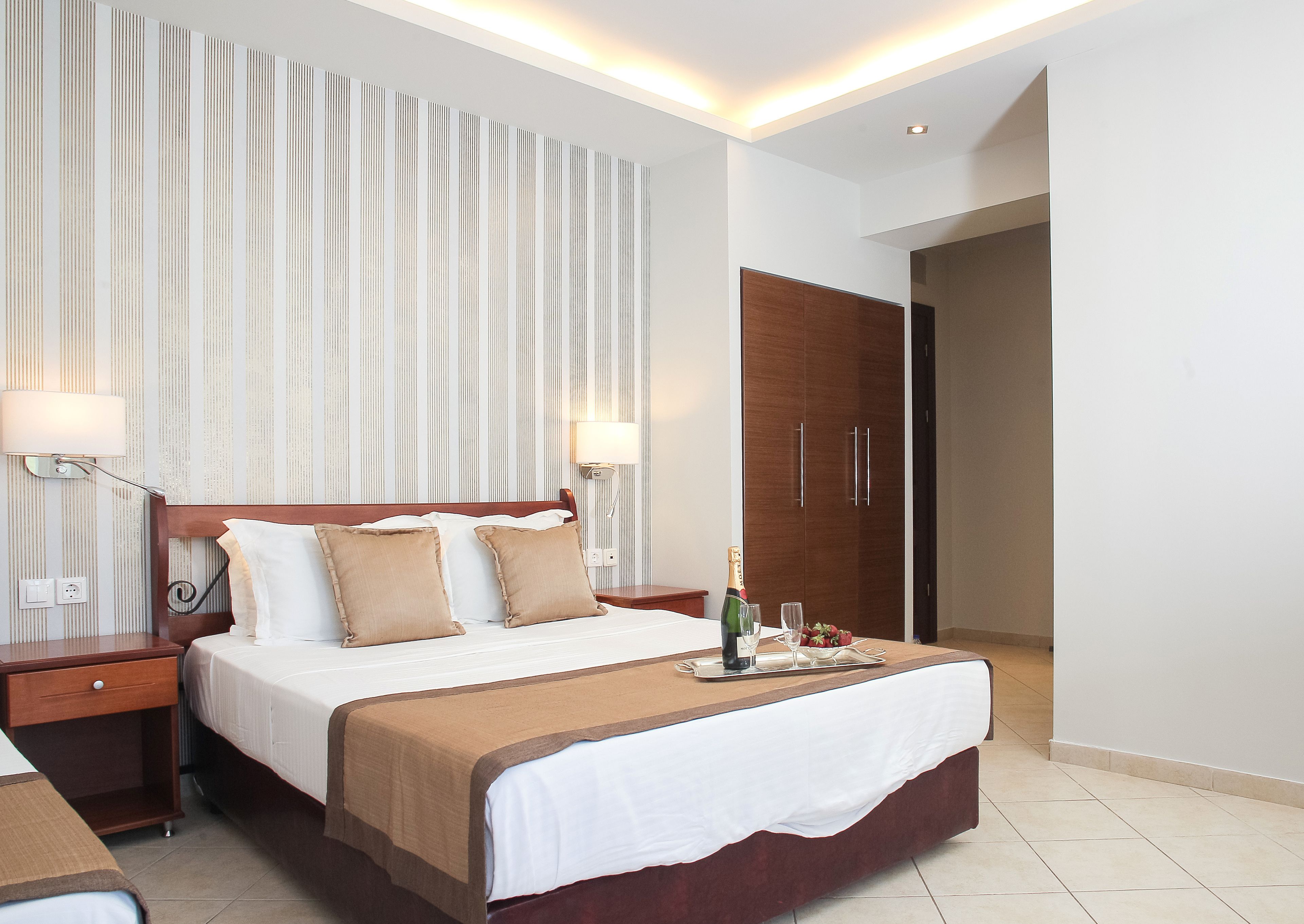 Habitación de hotel con una cama grande, cabecera de madera, dos lámparas de noche, mesita de noche, armario de madera y una bandeja con una botella y vasos sobre la cama.