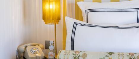 Una cama bien hecha con una colcha de motivos florales, una lámpara de noche, un teléfono antiguo y una botella de vino Panna sobre una mesita de noche.