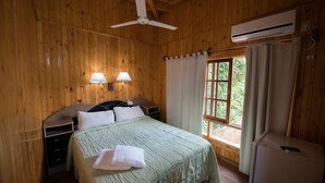 A wooden-paneled bedroom with a bed, two bedside lamps, a ceiling fan, an air conditioning unit, and a window with curtains. - Residencial Los Lapachos (Puerto Iguazú)