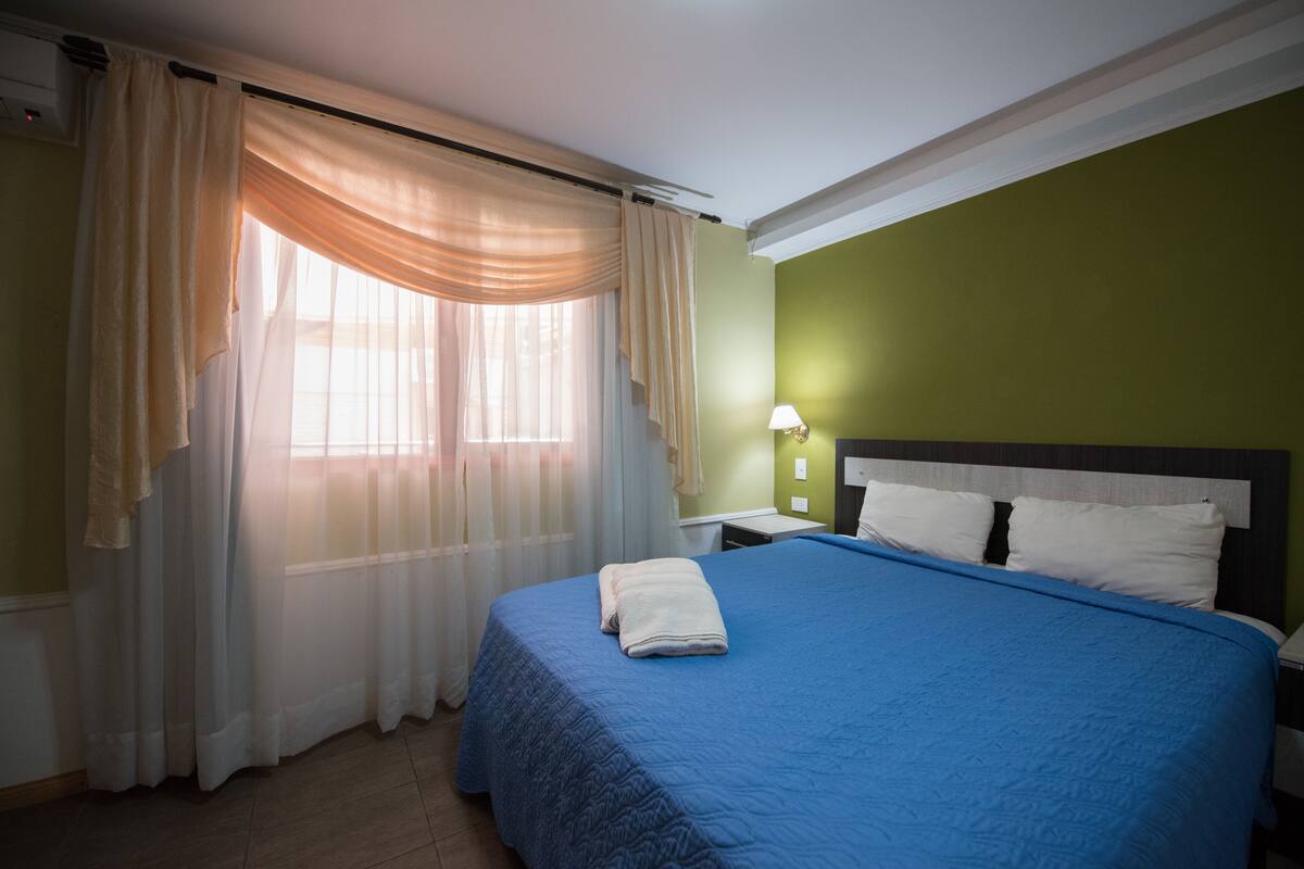 A neatly made bed with a blue bedspread, a white pillow, and a folded towel on the side. There is a nightstand with a lamp, and a window with sheer curtains allowing natural light.