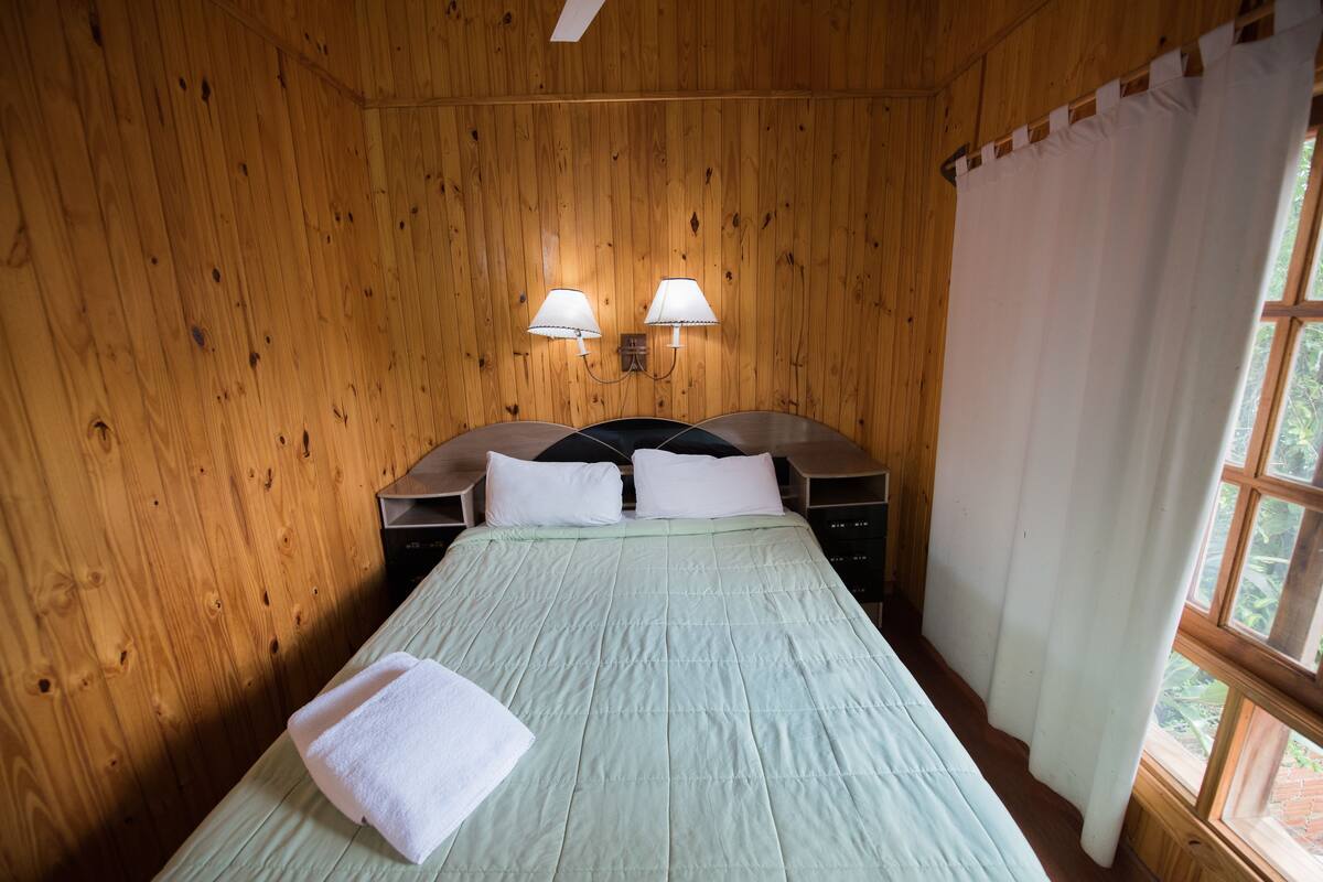 A wooden-paneled room with a bed, two bedside lamps, and a window with curtains.