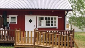 A red building with a white door and windows, a wooden deck, and a metal roof. - Pensionat Lövängen (Ljugarn)