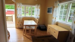A room with a wooden table and chairs, a small wooden cabinet, and a window with curtains. - Pensionat Lövängen (Ljugarn)