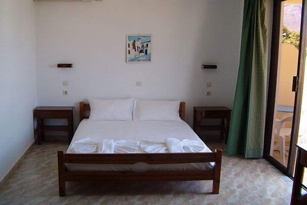 Un dormitorio con cama de madera, ropa de cama blanca, dos mesitas de noche, un cuadro en la pared y vistas a través de una puerta de cristal.