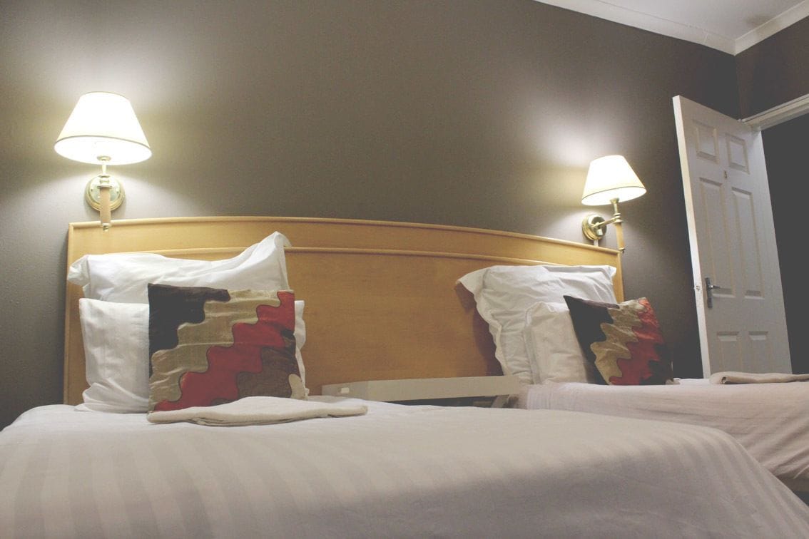 A hotel room with two beds, each with white bedding and decorative pillows. There are wall-mounted lamps on either side of the beds.