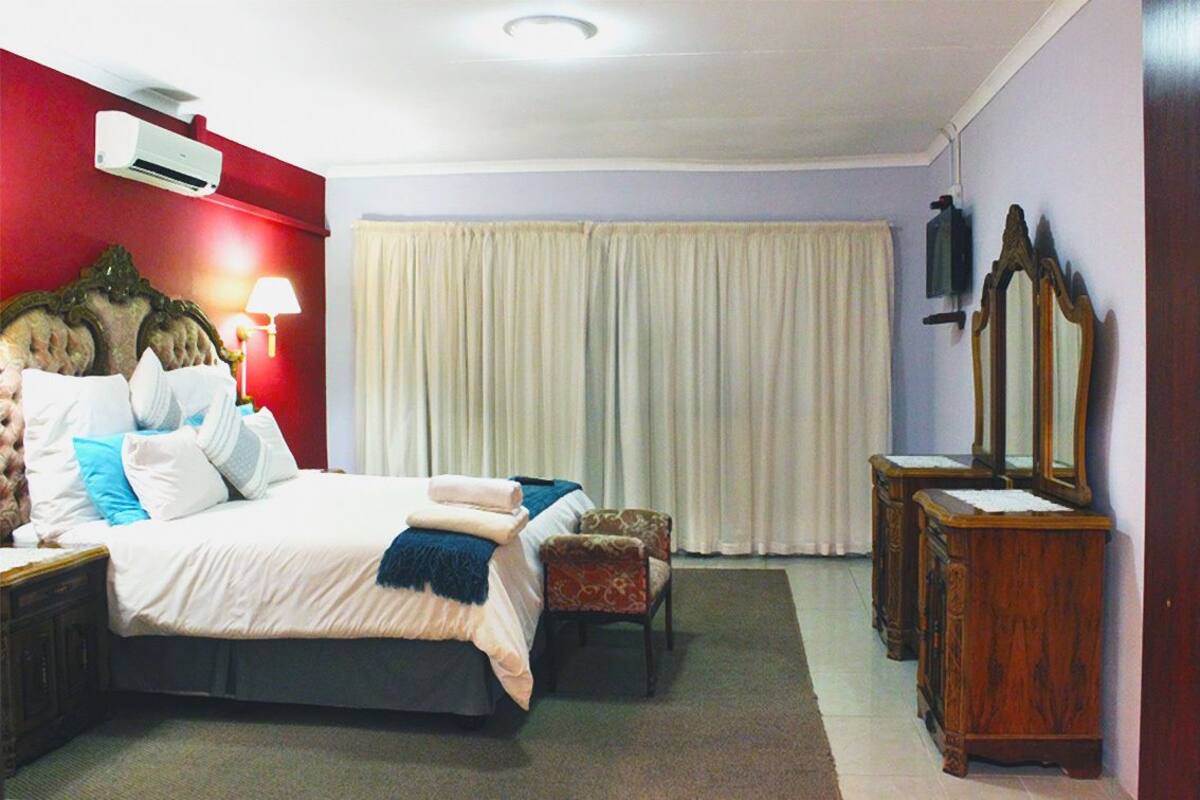 Ein Hotelzimmer mit einem großen Bett, einem Nachttisch, einem Sessel, einer Holzkommode, einem Spiegel und einem Fenster mit Vorhängen.