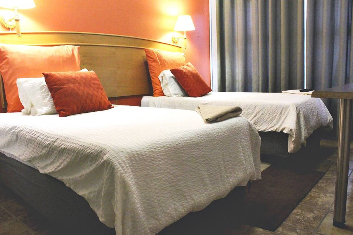 Ein Hotelzimmer mit zwei Betten, jeweils mit weißer Bettdecke und mehreren Kissen, darunter orangefarbene und weiße. An beiden Seiten der Betten befinden sich jeweils eine Wandlampe und ein Fenster mit Vorhängen.