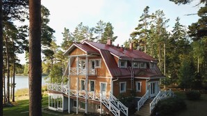 Punakattoinen, valkoisilla kaiteilla varustettu kaksikerroksinen puutalo, jonka edessä on laituri, joka ulottuu järvelle.