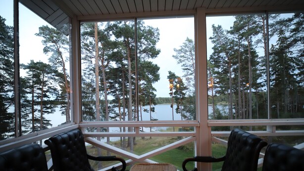 Katettu ulkoalue, jossa on kaksi mustaa nahkaista tuolia ja punottu pöytä. Alueelta avautuu näkymä järvelle ja metsäiselle alueelle.
