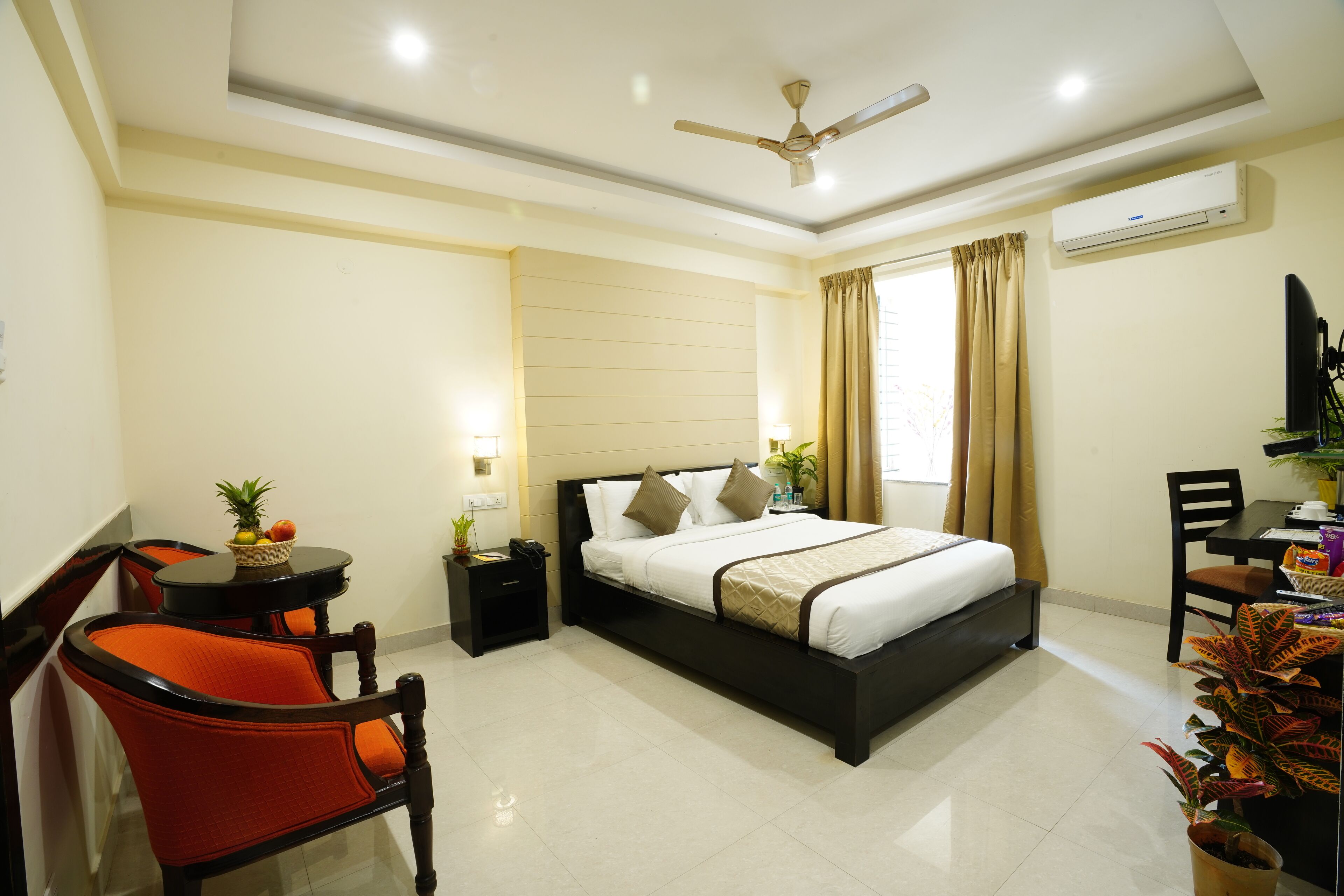 A hotel room with a bed, a desk, a chair, a small table with a fruit bowl, and a potted plant.