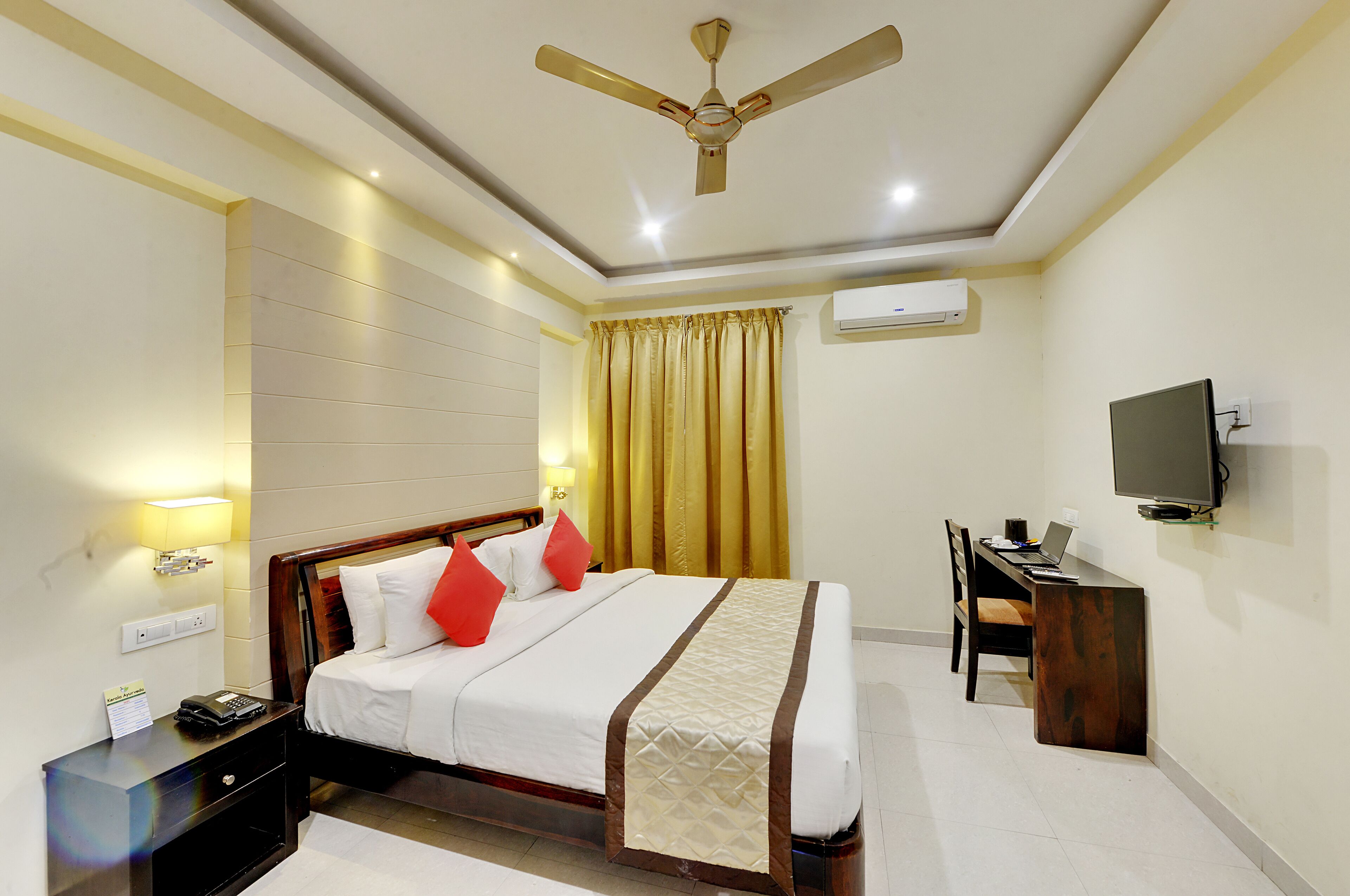 A hotel room with a bed, bedside table, desk, chair, TV, and ceiling fan.