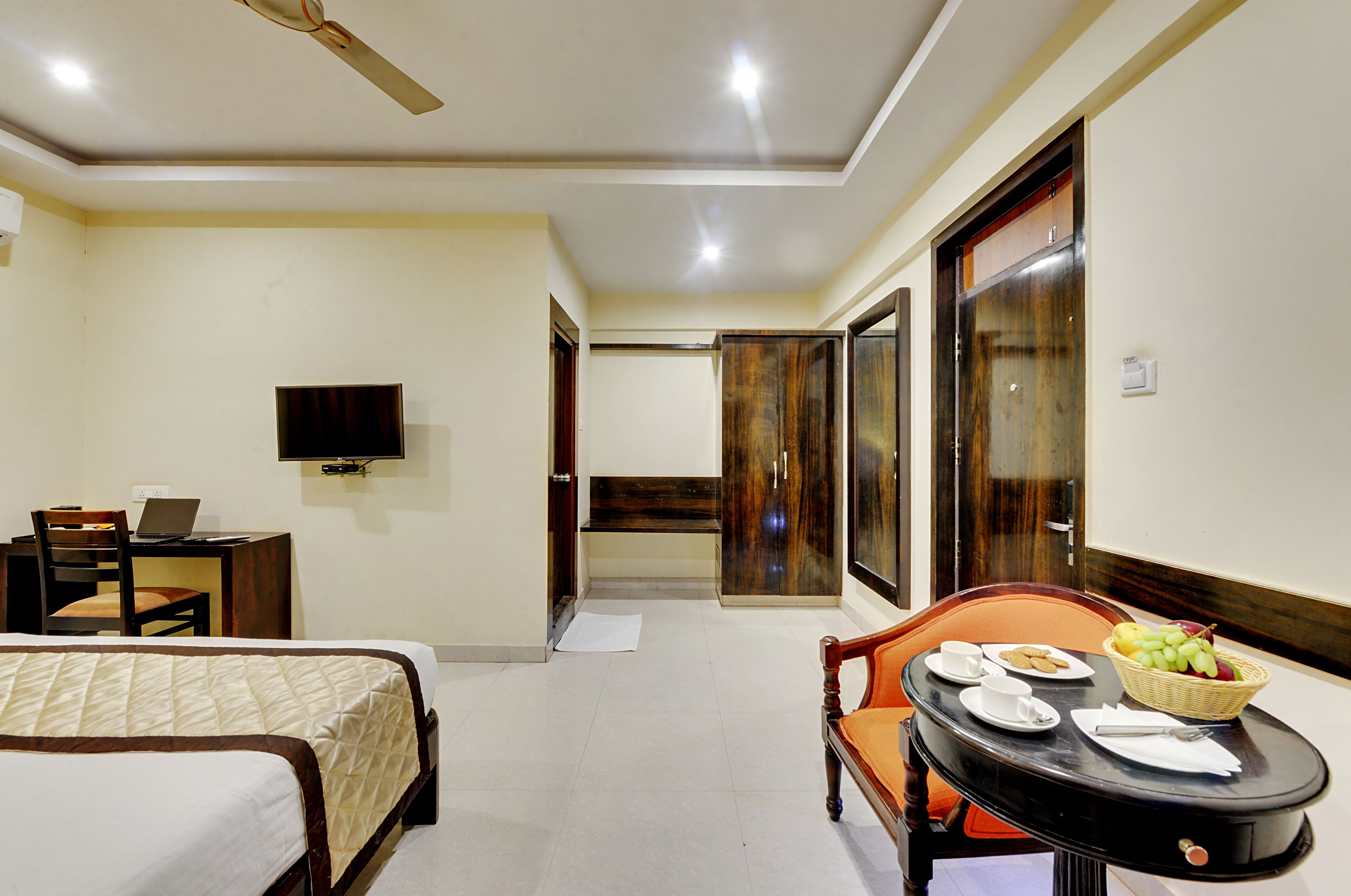 Une chambre d’hôtel comprenant un lit, un bureau avec un ordinateur portable, une télévision et un coin repas avec des assiettes et un panier de fruits.