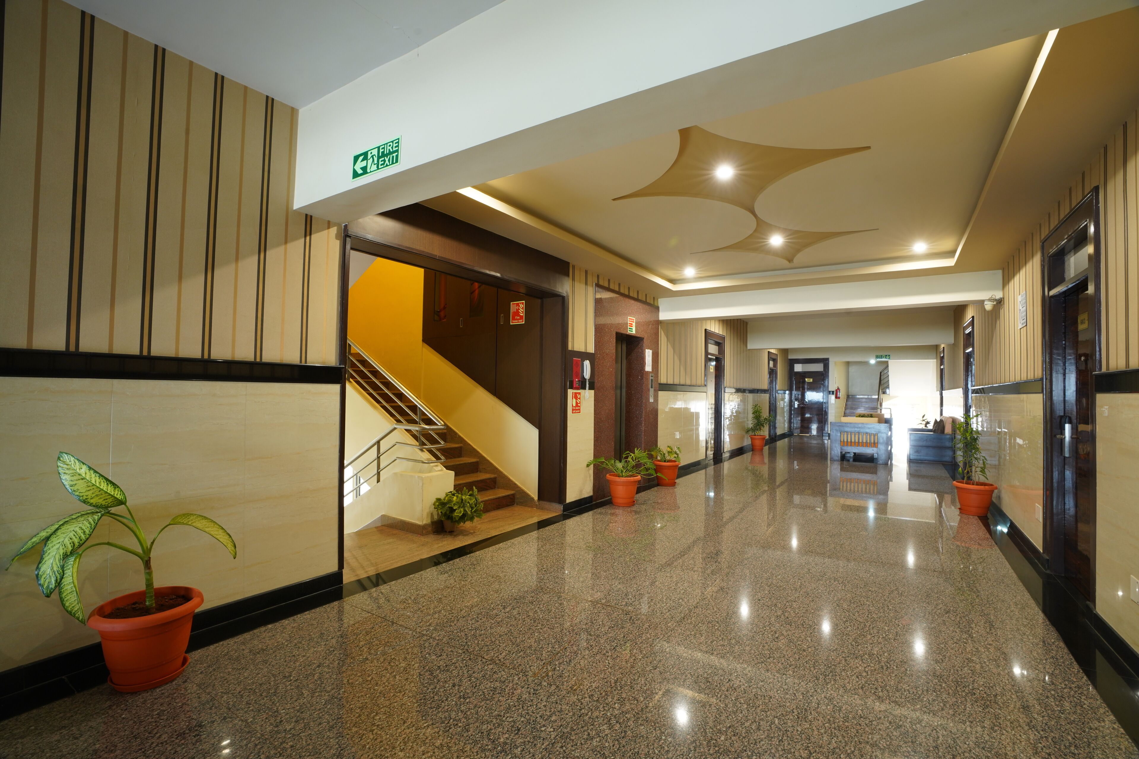 Pasillo de hotel con piso pulido, plantas en macetas, una escalera y señal de salida de incendios.