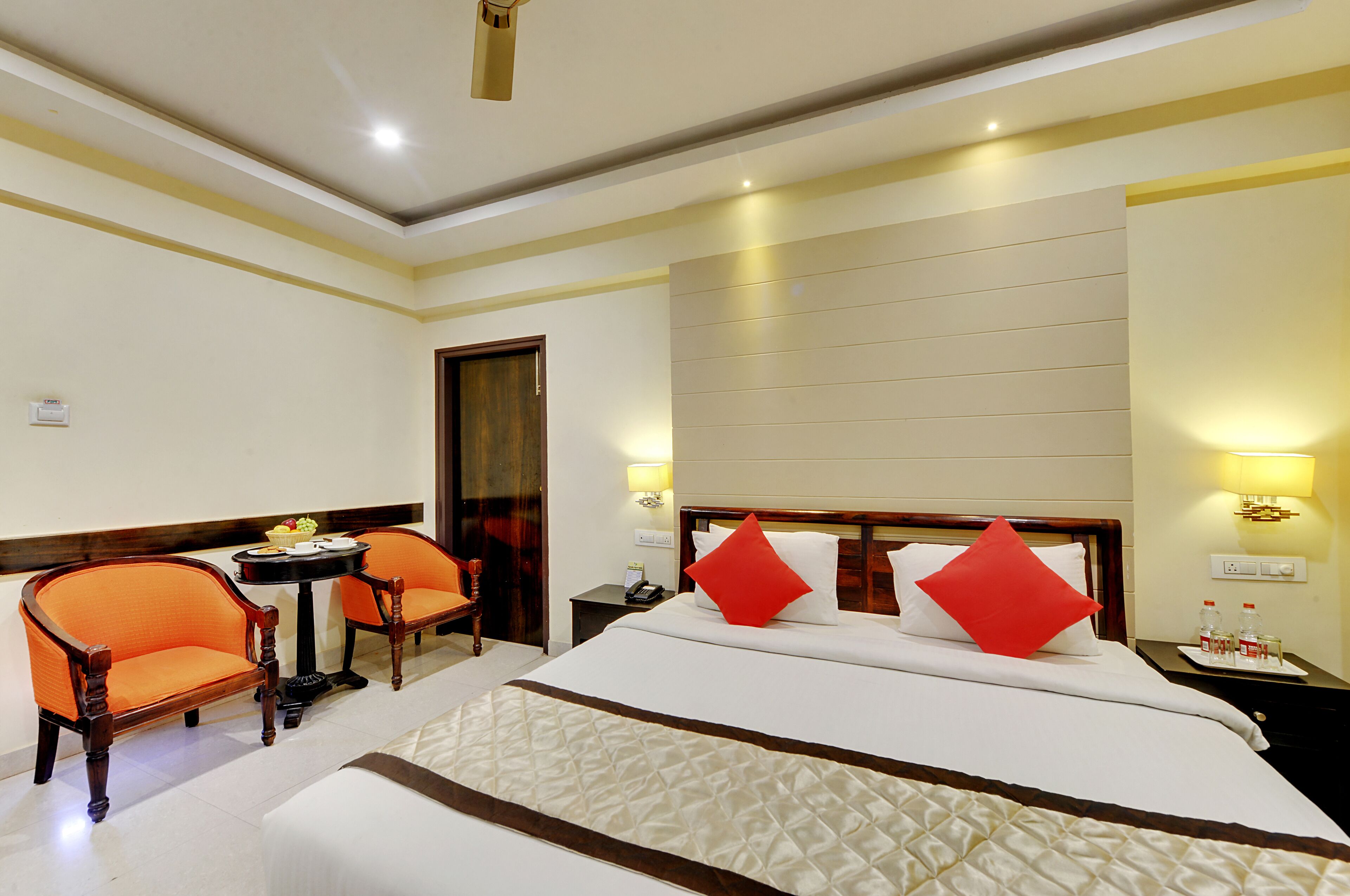 A hotel room with a large bed, two orange chairs, a small table, and a nightstand with a lamp.