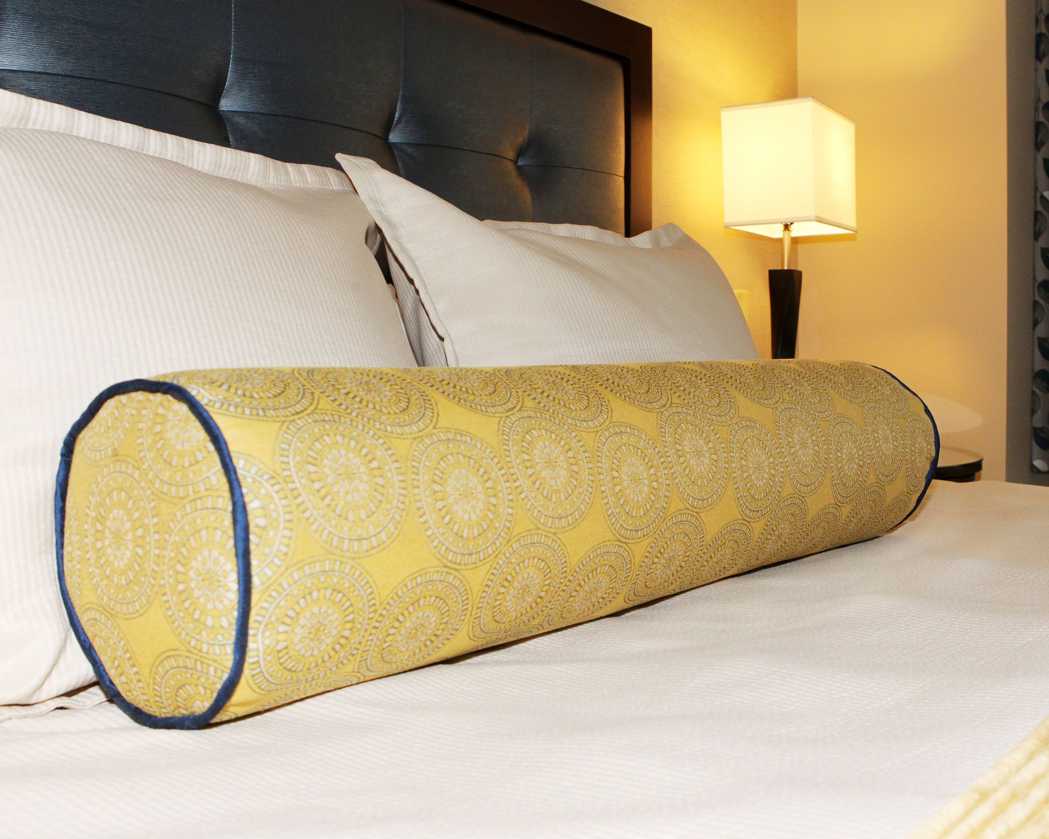 A bed with a rolled-up decorative pillow, a headboard, and a bedside lamp.