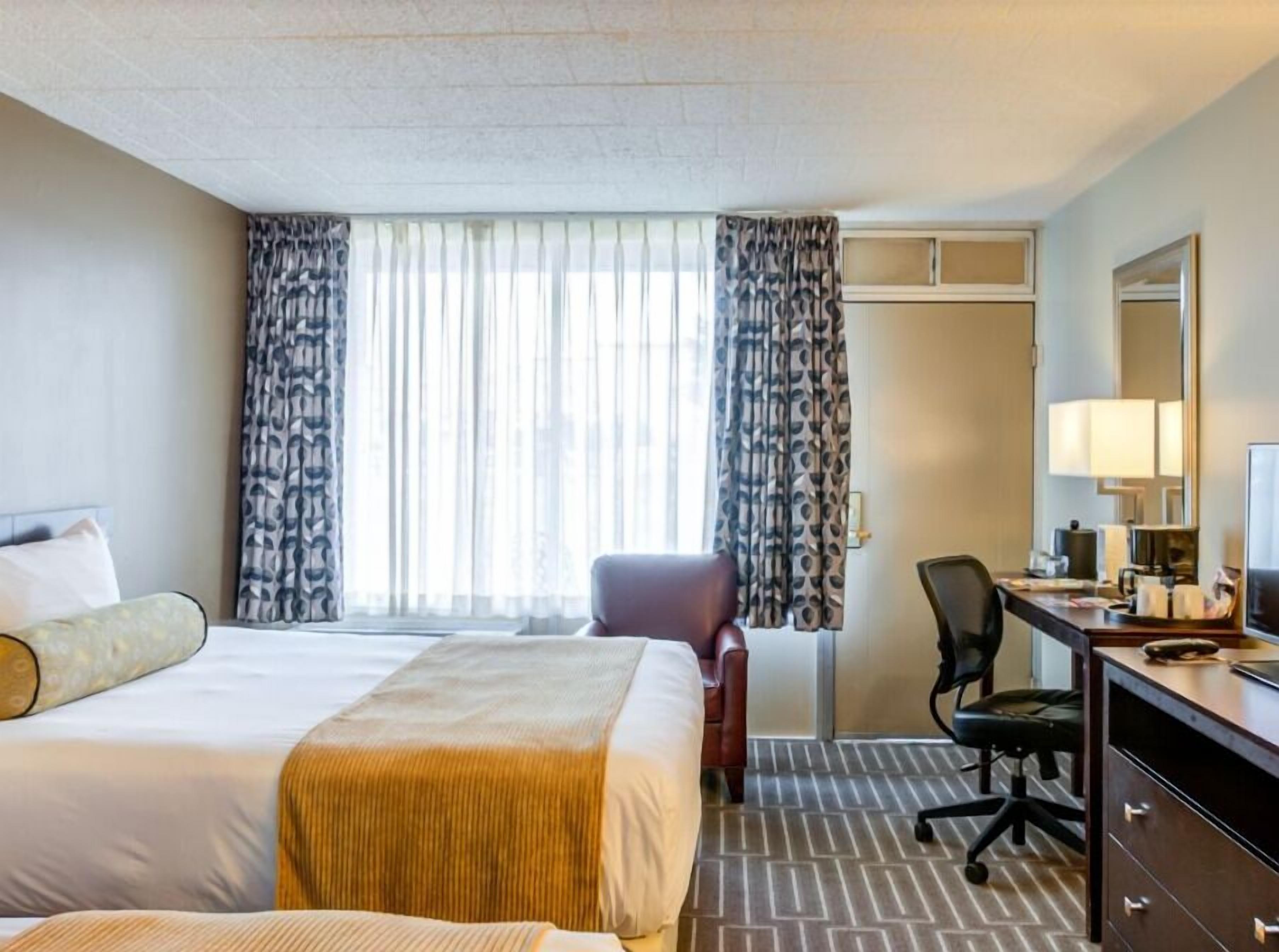 Une chambre d’hôtel avec un lit, un bureau, une chaise, un miroir et une fenêtre avec des rideaux.