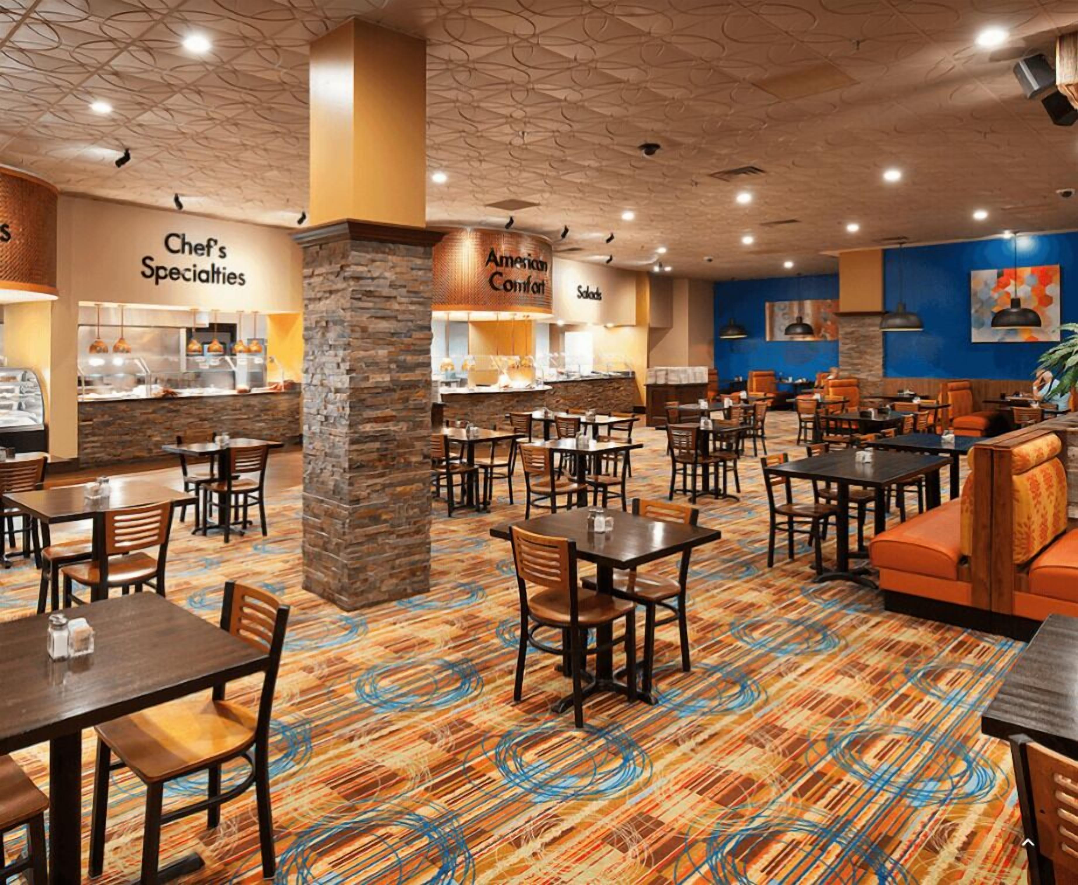 A hotel lobby with multiple dining tables and chairs, a kitchen area labeled 'Chef's Specialties', and an 'American Comfort' sign.