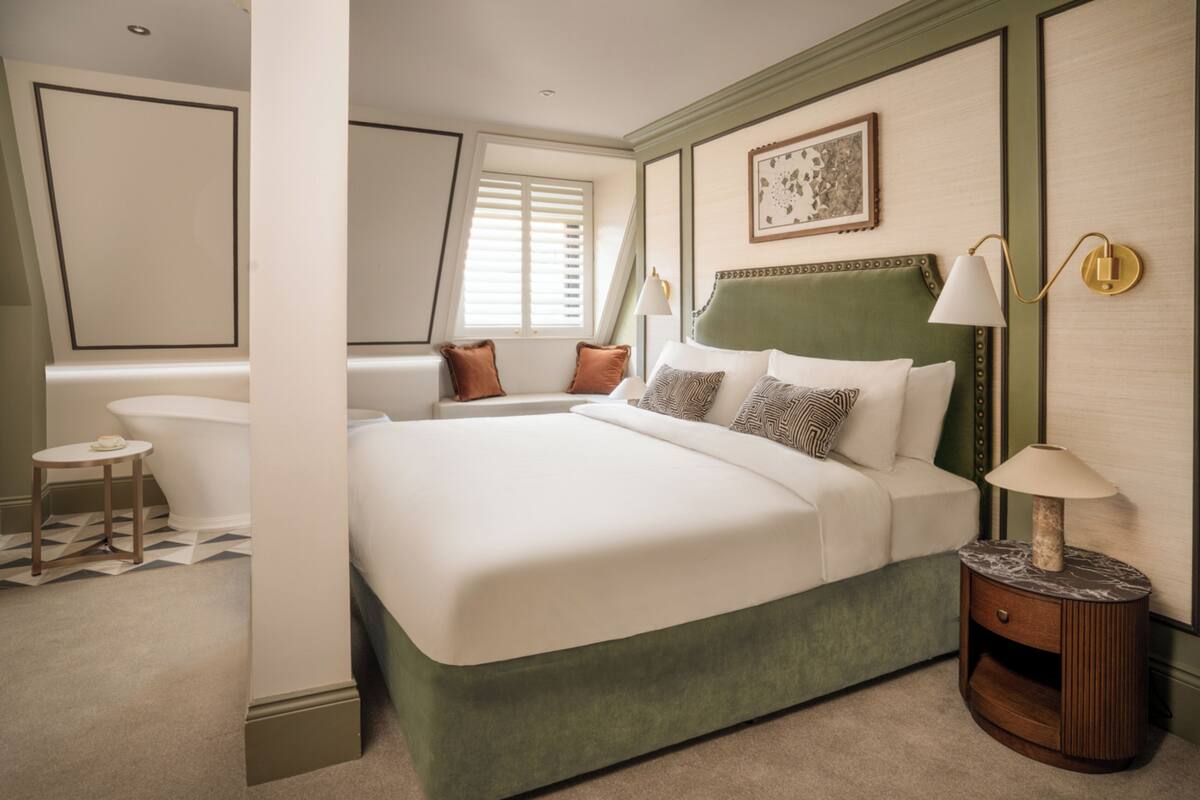 A hotel room with a large bed, a small round table, a bathtub, and a window with shutters.