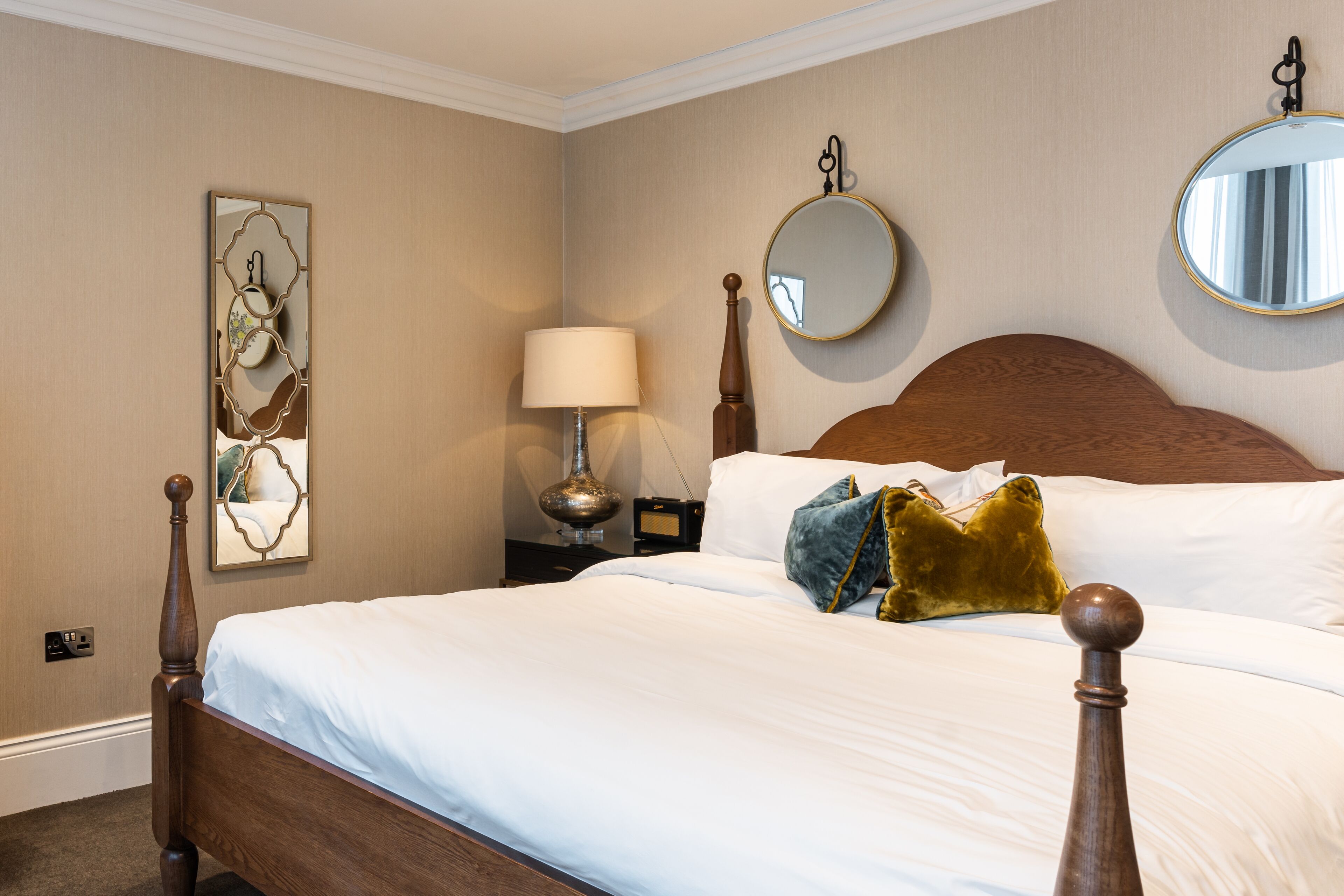 A neatly made bed with white linens, a wooden headboard, and decorative pillows. There is a round mirror on the wall and a lamp on the nightstand.