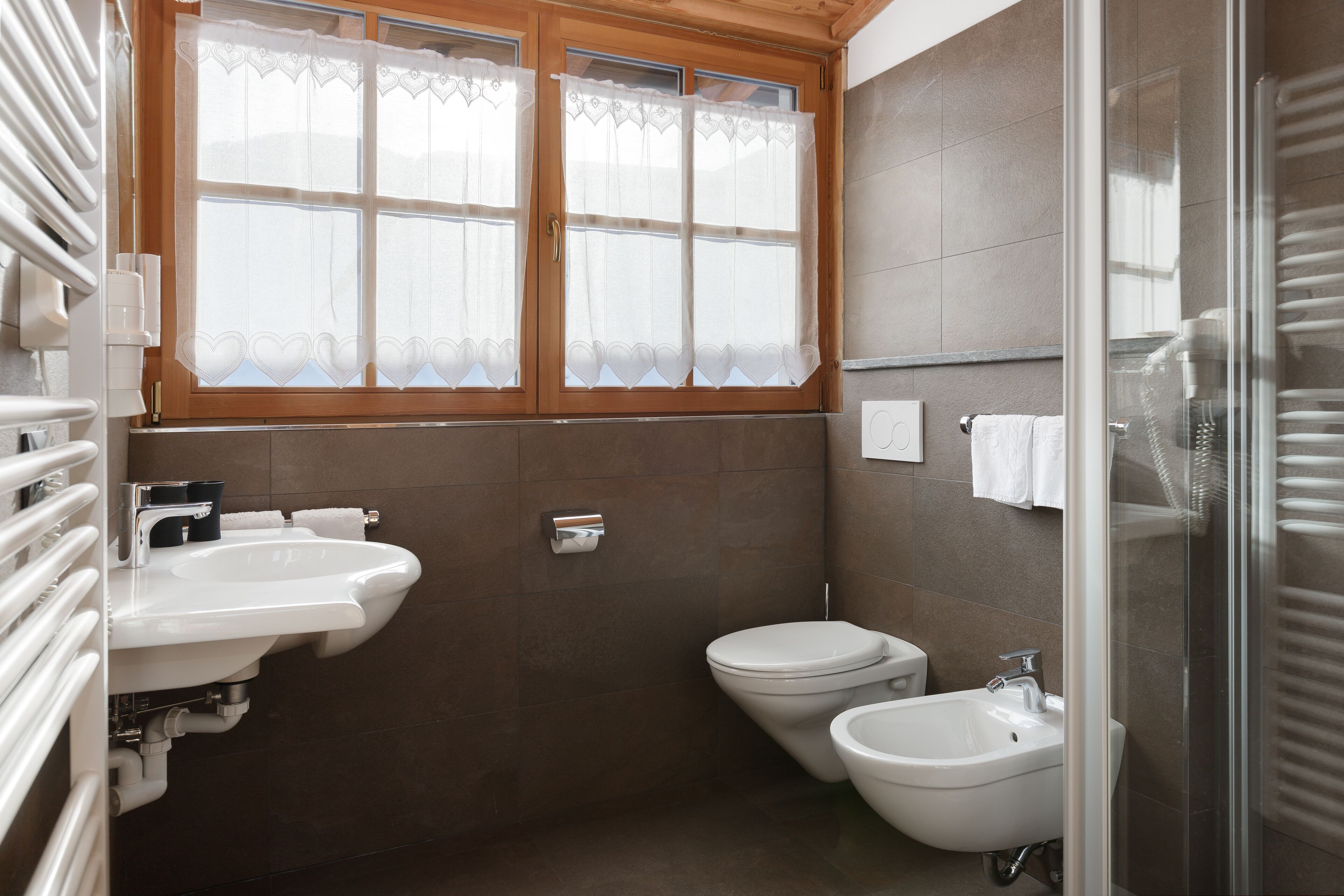 A bathroom with a sink, bidet, toilet, and a window with lace curtains.