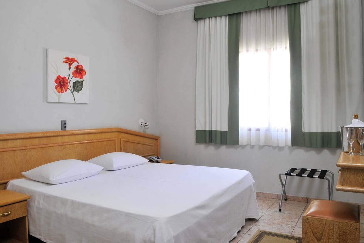 Une chambre d’hôtel avec un lit, une table de chevet, une petite table avec une lampe, une fenêtre avec des rideaux et un tableau au mur.