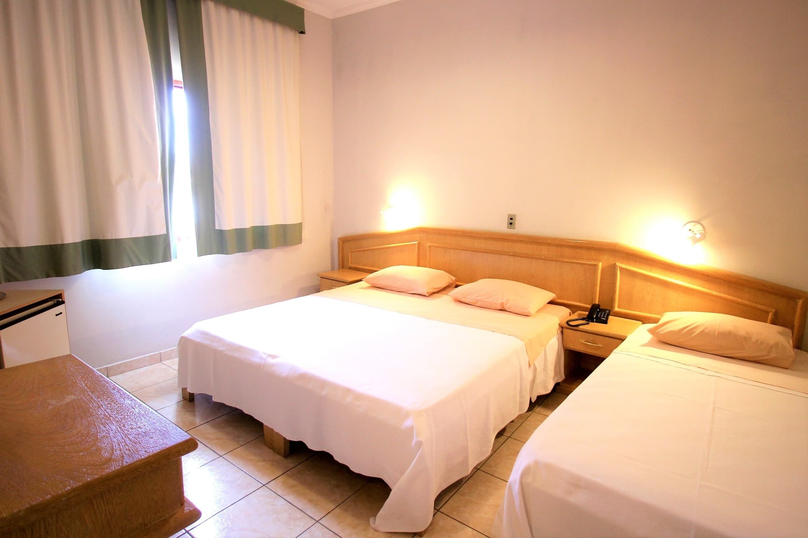 A hotel room with two beds, a wooden nightstand, a desk, and a window with curtains.
