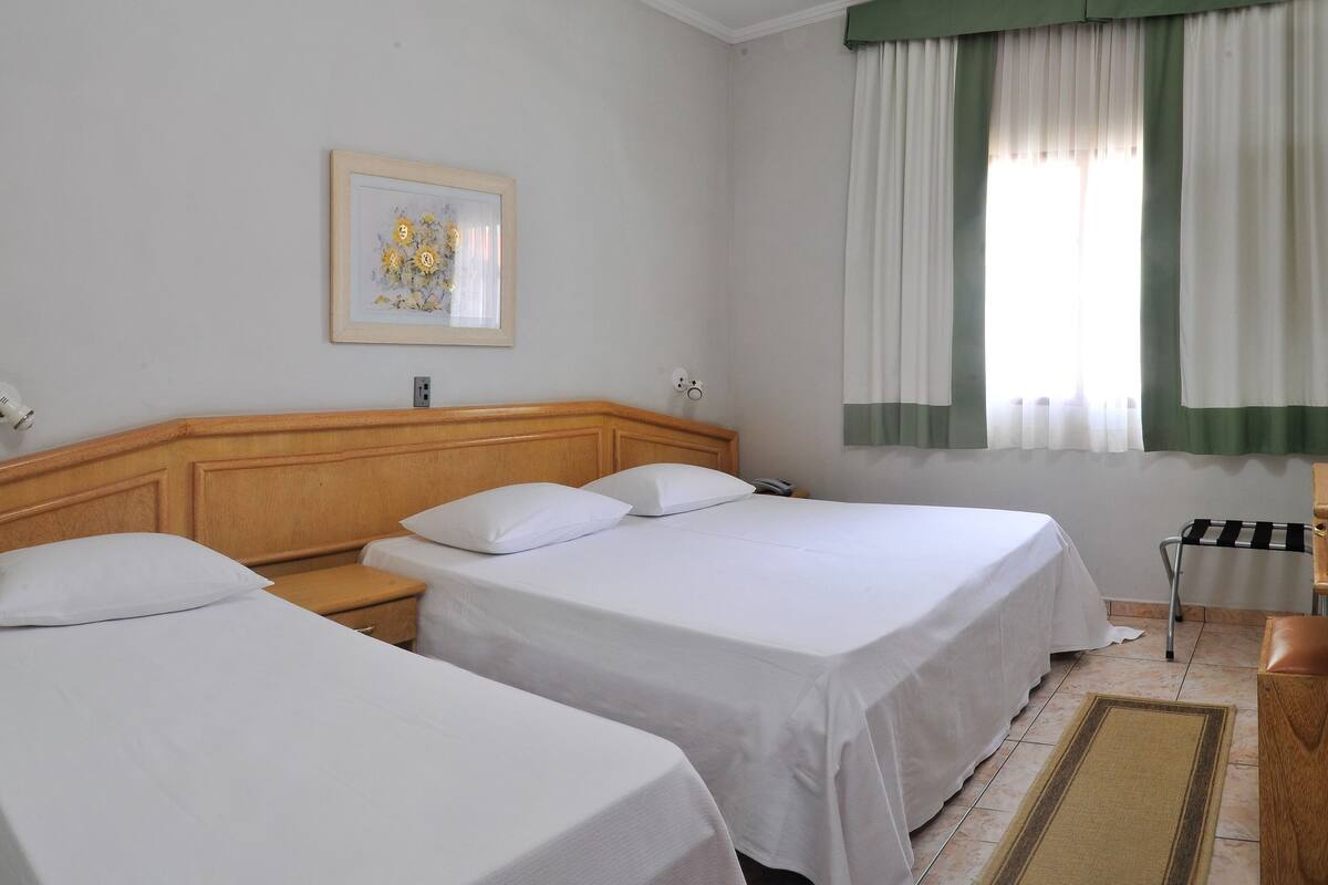 Une chambre d’hôtel avec deux lits simples, une tête de lit en bois, une table de chevet avec une lampe, un tableau encadré au mur, une fenêtre avec des rideaux verts et une commode en bois.