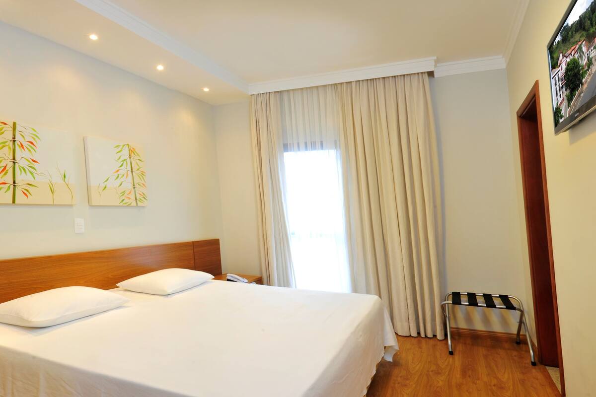 Une chambre d’hôtel avec un grand lit, une table de chevet, une petite table avec un téléphone, une fenêtre avec des rideaux et un téléviseur fixé au mur.