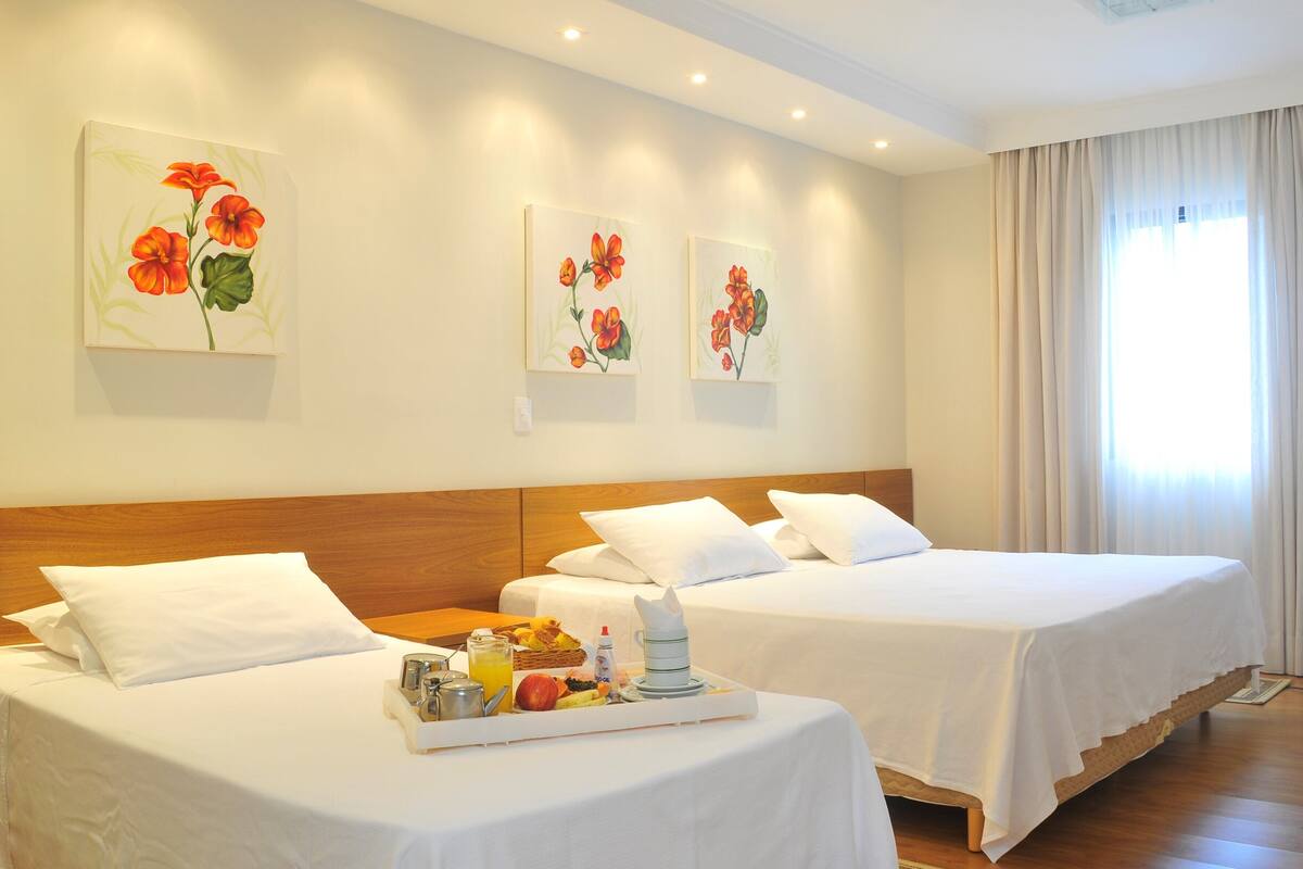 Une chambre d’hôtel avec deux lits, du linge de lit blanc, un plateau de petit-déjeuner avec des fruits et une boisson, et une décoration florale au mur.