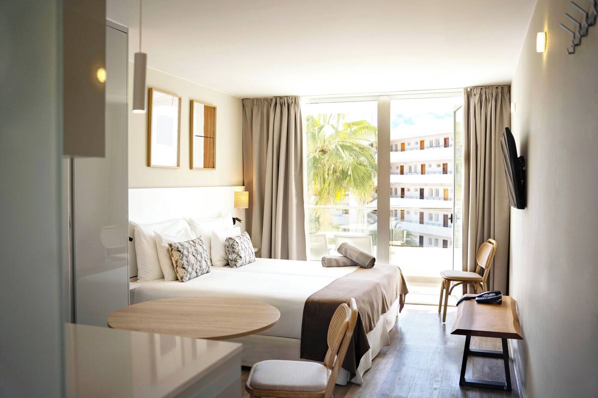Une chambre d’hôtel avec un lit, une petite table, une chaise, une télévision et un balcon offrant une vue sur les immeubles et les palmiers.