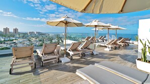 A rooftop lounge area with sun loungers and umbrellas, overlooking a cityscape and the sea.