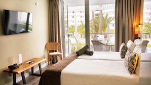A hotel room with two beds, a TV, a desk, and a balcony with a view of palm trees and buildings.
