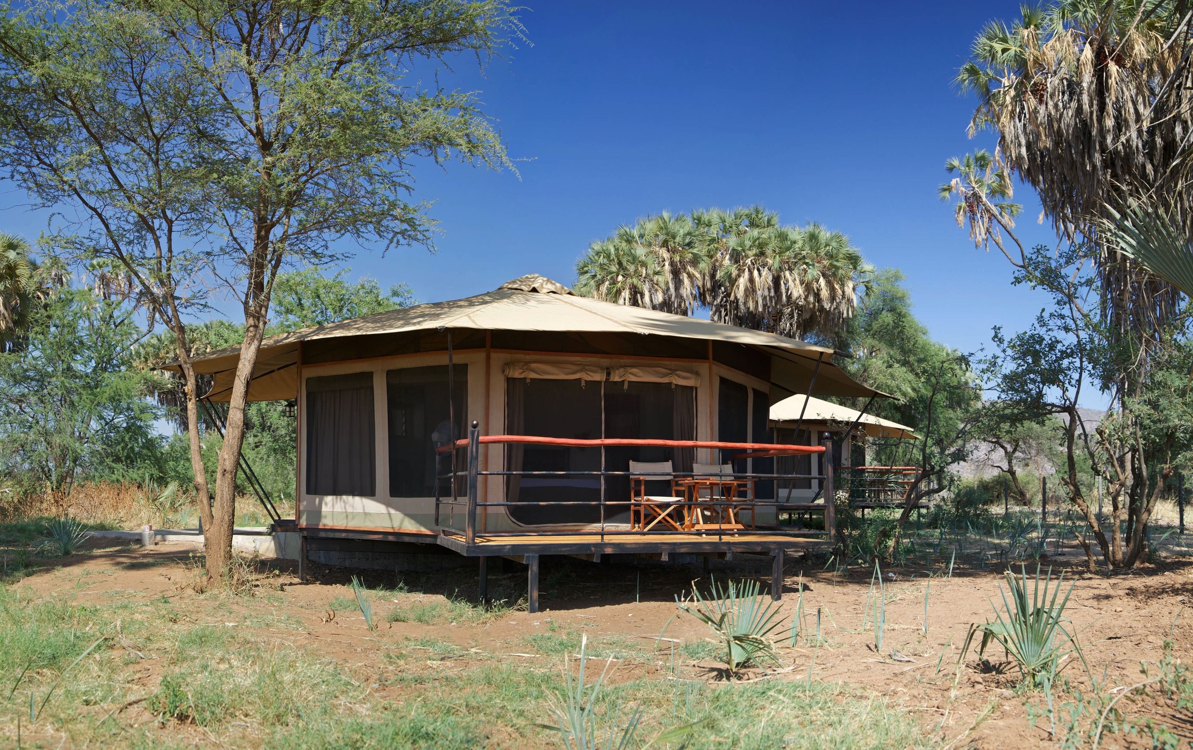 A safari lodge with a covered veranda, outdoor seating, and a dirt ground.
