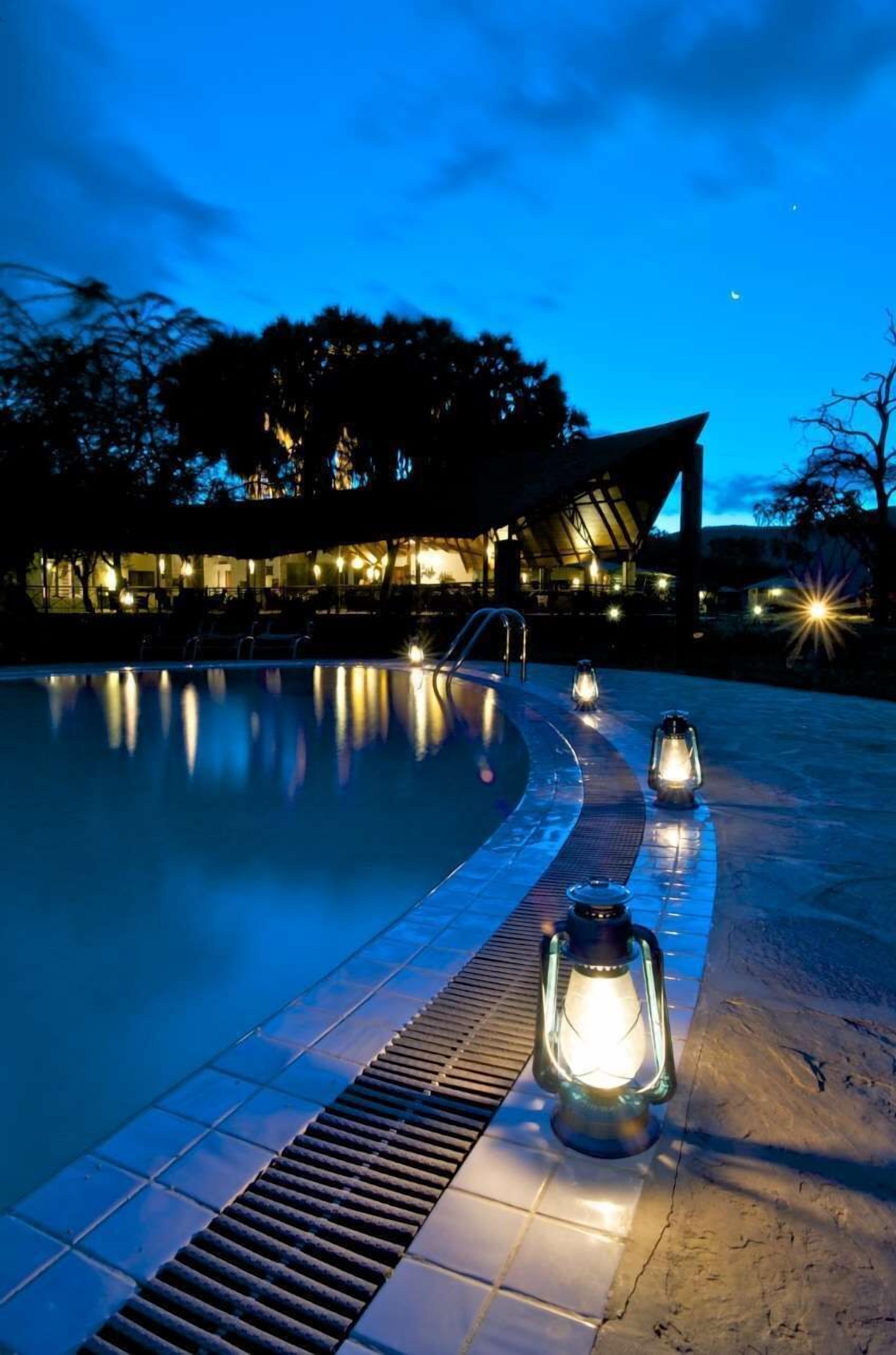 A swimming pool at dusk with lanterns along the edge, a covered seating area, and a building in the background.
