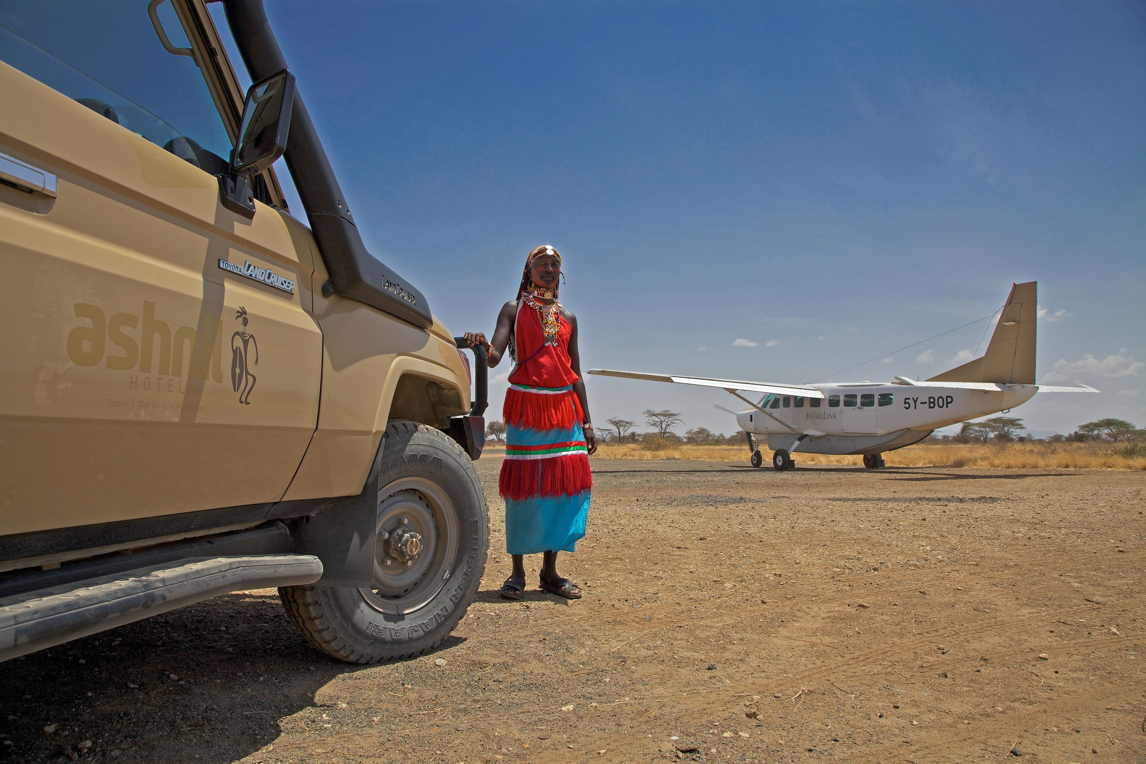 Una persona de pie junto a un vehículo de safari y un pequeño avión en un paisaje seco y abierto.