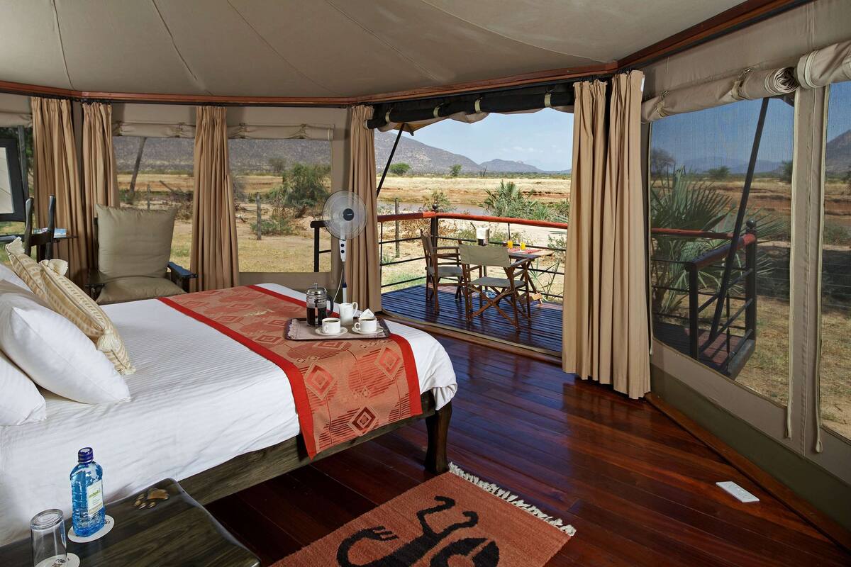 A bedroom with a bed, a table with a bottle and glasses, a fan, and a view of the outdoors.