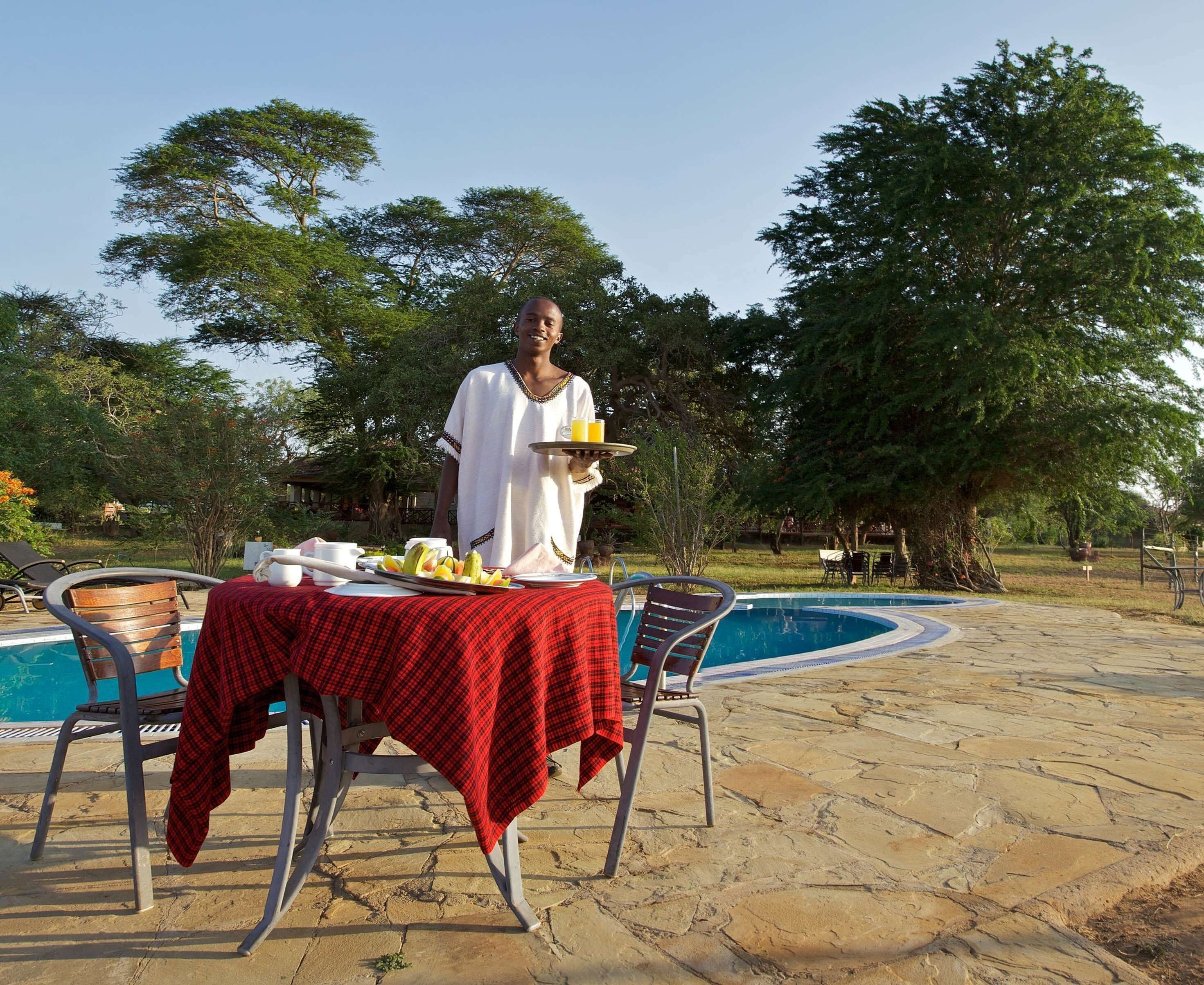 A dining area with a table set for two, a pool in the background, and a waiter serving a tray.