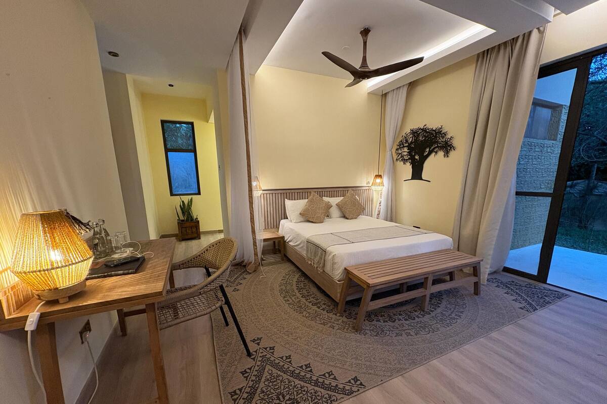 A bedroom with a bed, bedside table, lamp, chair, and a view of the outdoors.
