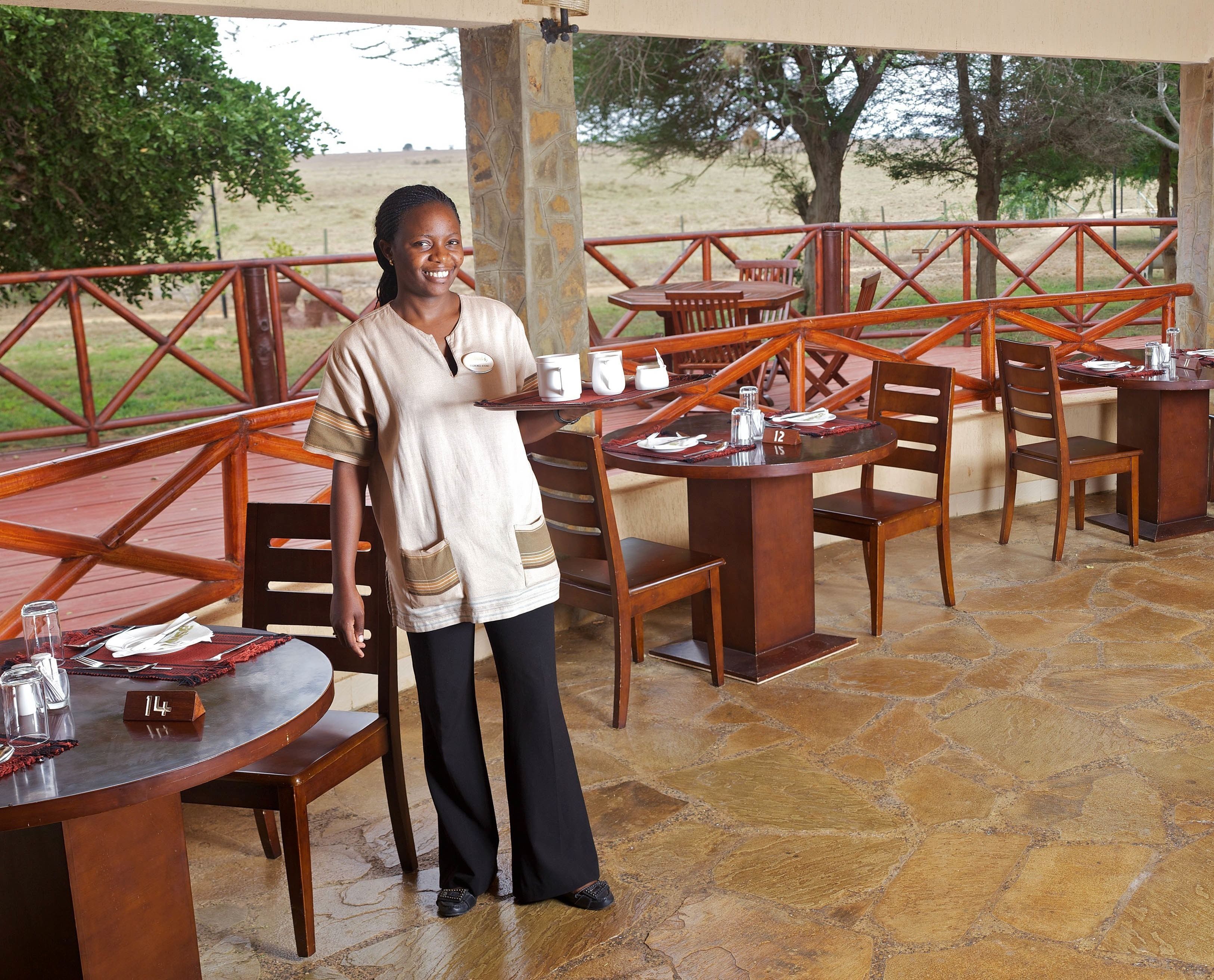 A person standing in an outdoor dining area with stone flooring and wooden furniture. Tables are numbered, and there are chairs and a bench.