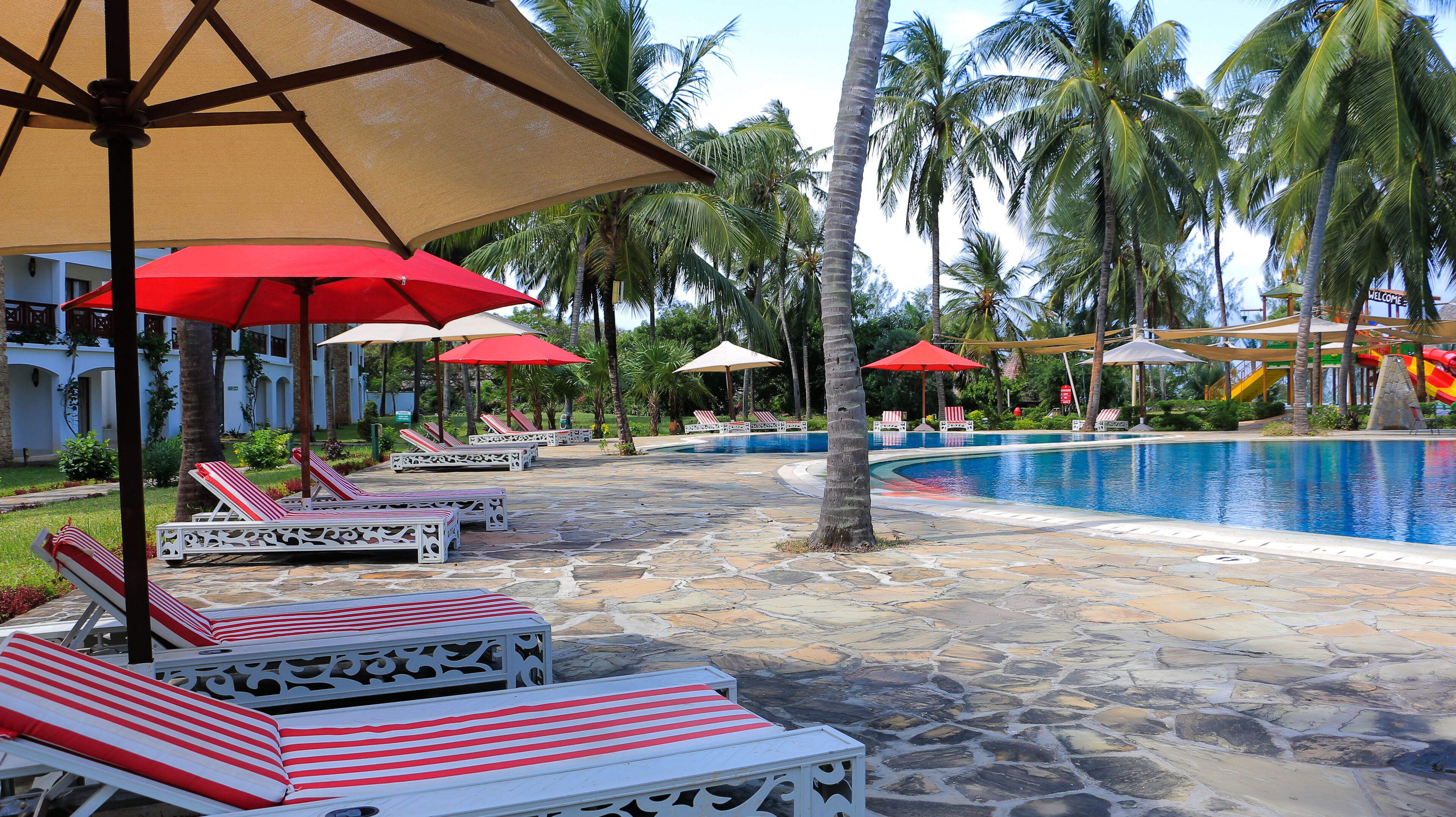 A pool area with lounge chairs and umbrellas, surrounded by palm trees and a building with a 'Welcome' sign.