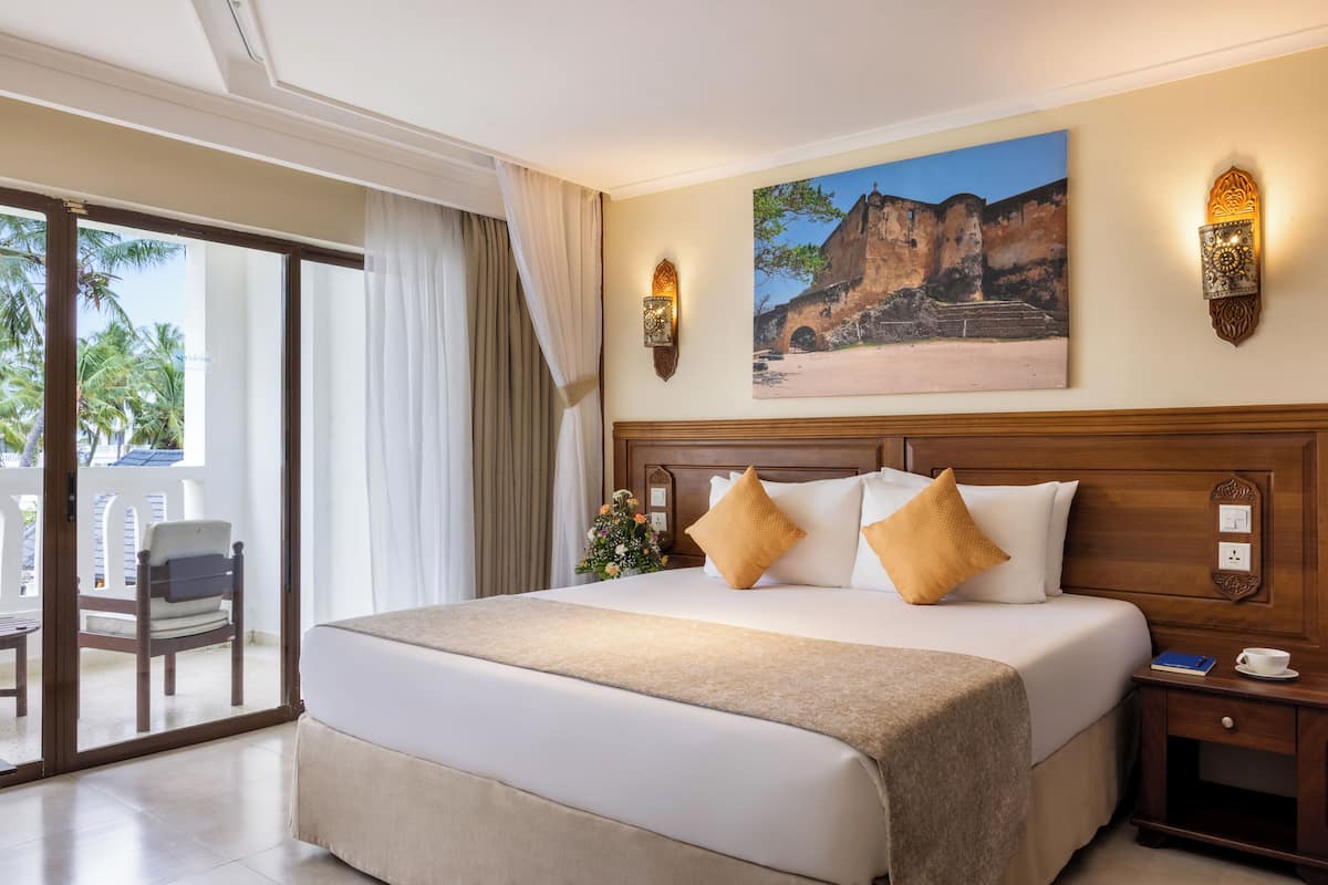 Habitación de hotel con una cama grande, una mesita de noche, una silla, un balcón con vistas a palmeras y un cuadro en la pared.