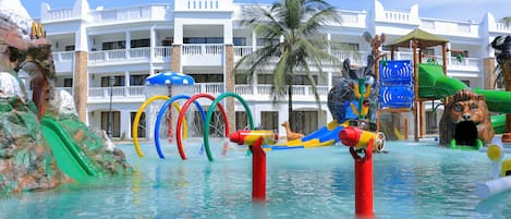 A water park with slides and play structures, surrounded by a hotel with multiple balconies and palm trees.