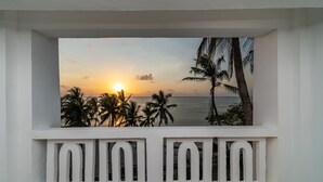 Uma varanda com vista para o pôr do sol sobre o oceano, palmeiras e um corrimão branco.