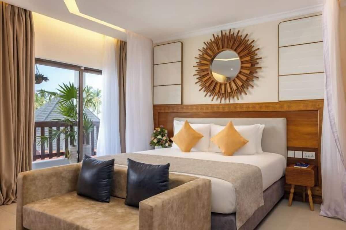 Habitación de hotel con cama, sofá, mesita de noche, espejo y vista a palmeras.